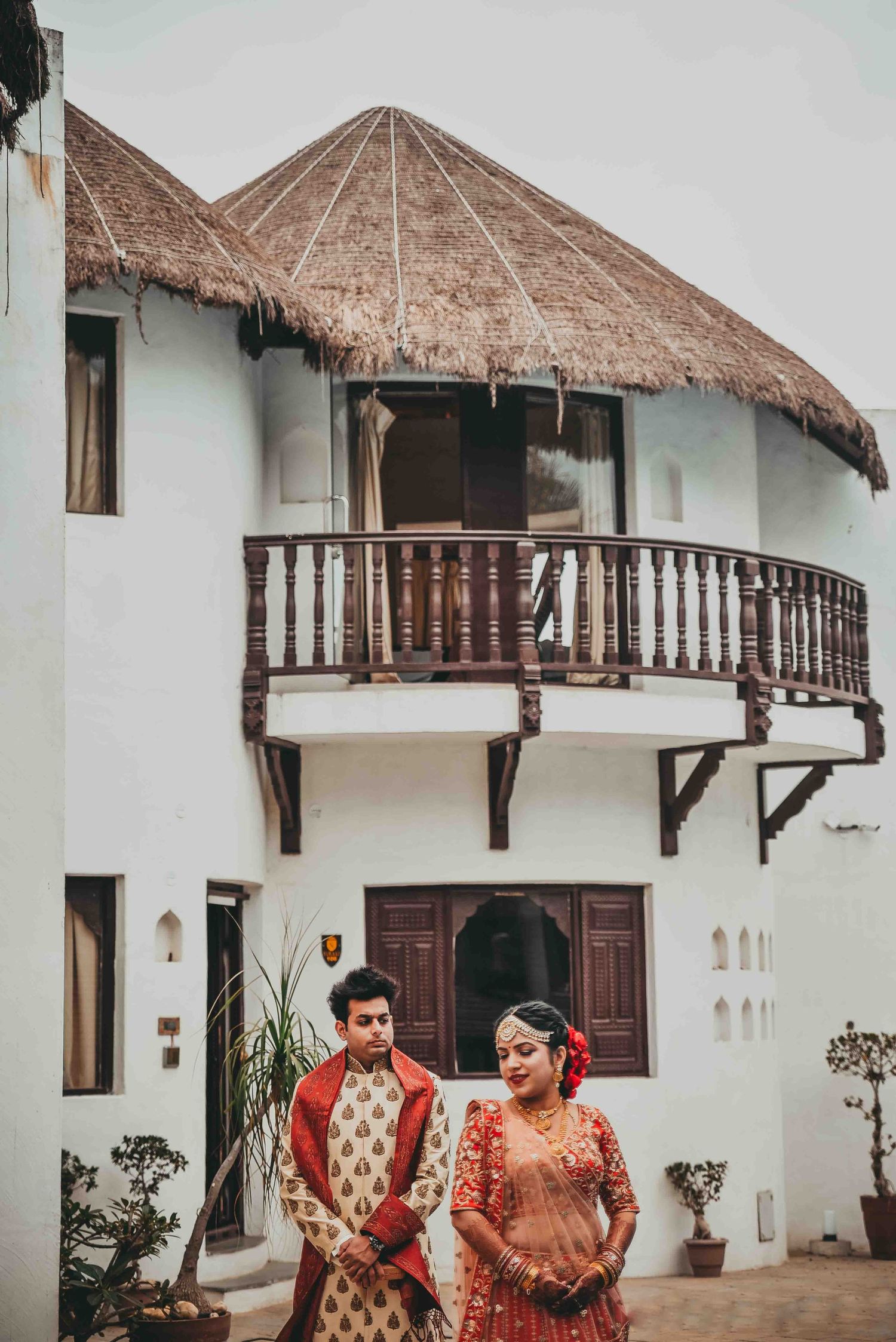 Indian bride and groom in traditional red and gold outfits standing before a white resort with thatched roof balcony.