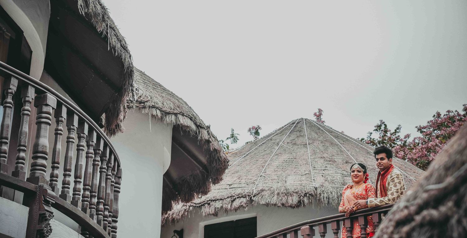 Indian wedding couple in traditional attire posing on a balcony of a resort with thatched roof bungalows.