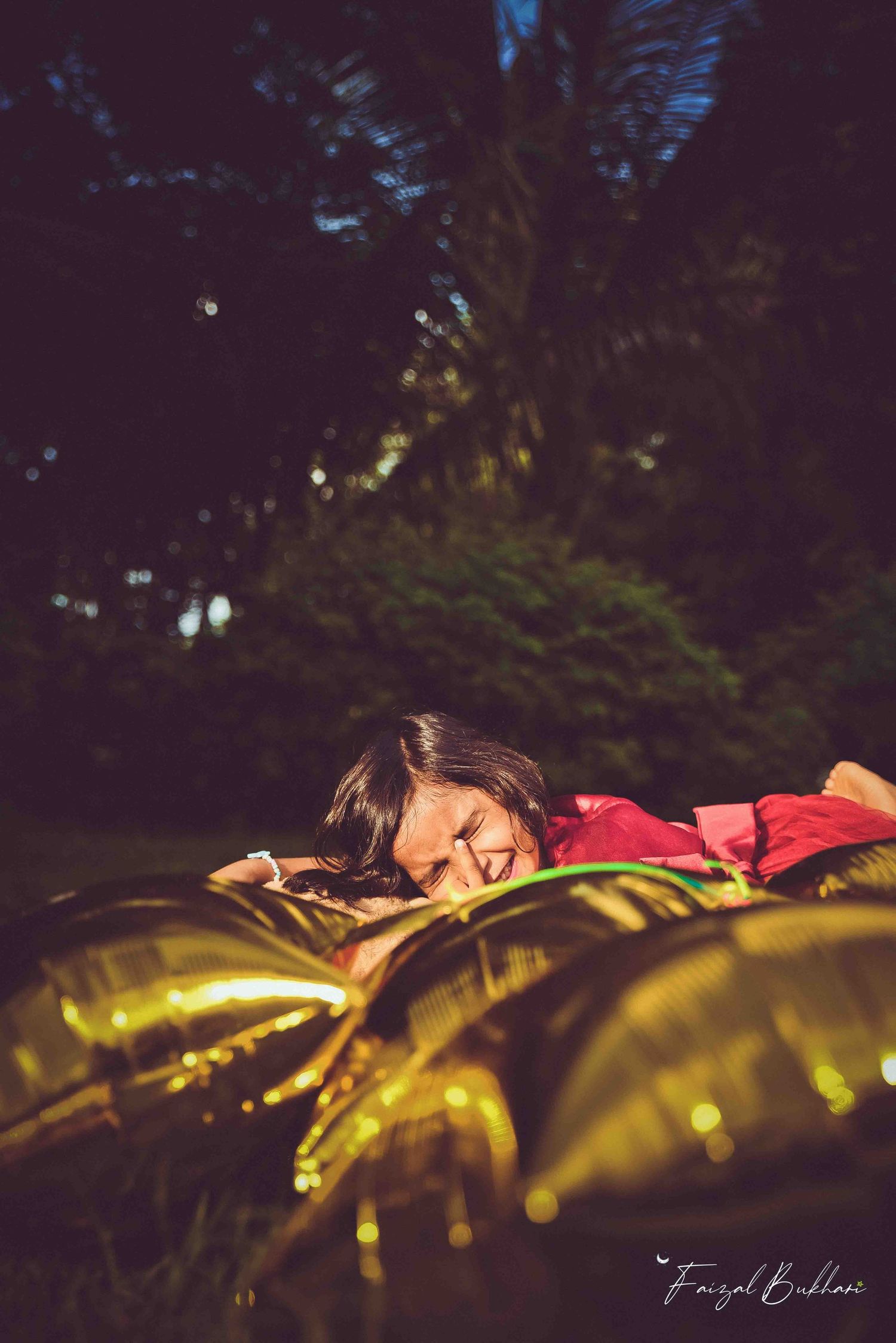 A young girl in red rests among gold star balloons on grass in a softly lit garden setting.