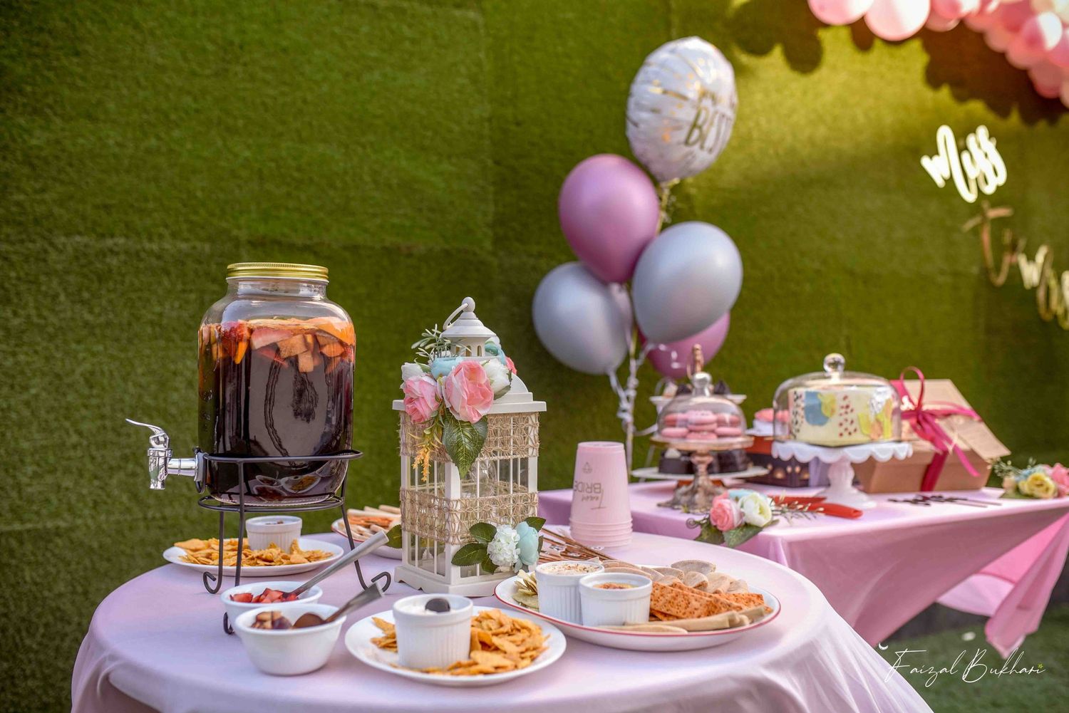 Pink bridal shower dessert table with large beverage dispenser, balloons, cake, and assorted snacks outdoors.