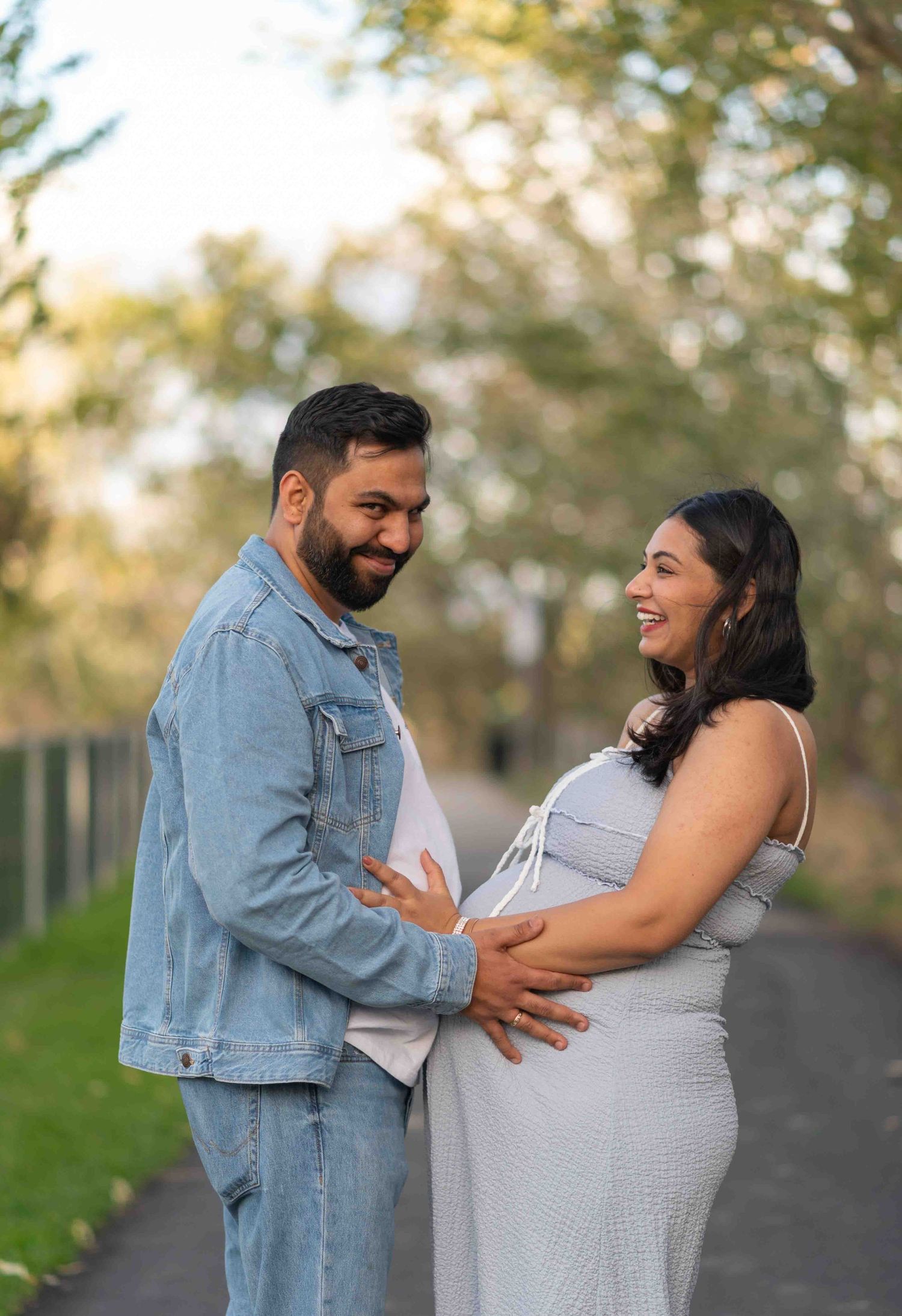 Smiling couple cradling pregnant belly together on tree-lined path during golden hour.