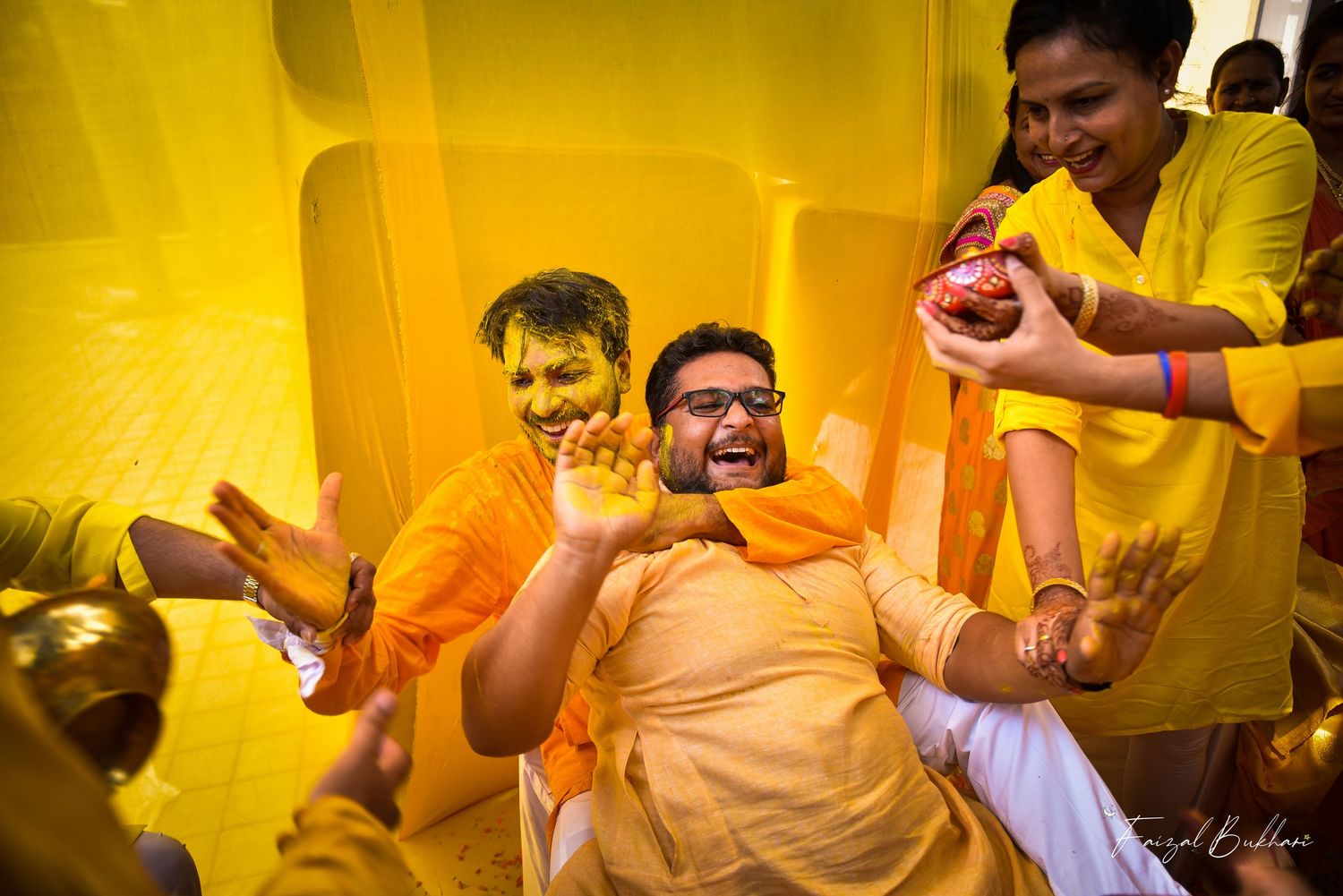 Joyful Indian groom laughing during colorful Haldi ceremony surrounded by family and friends in yellow outfits.