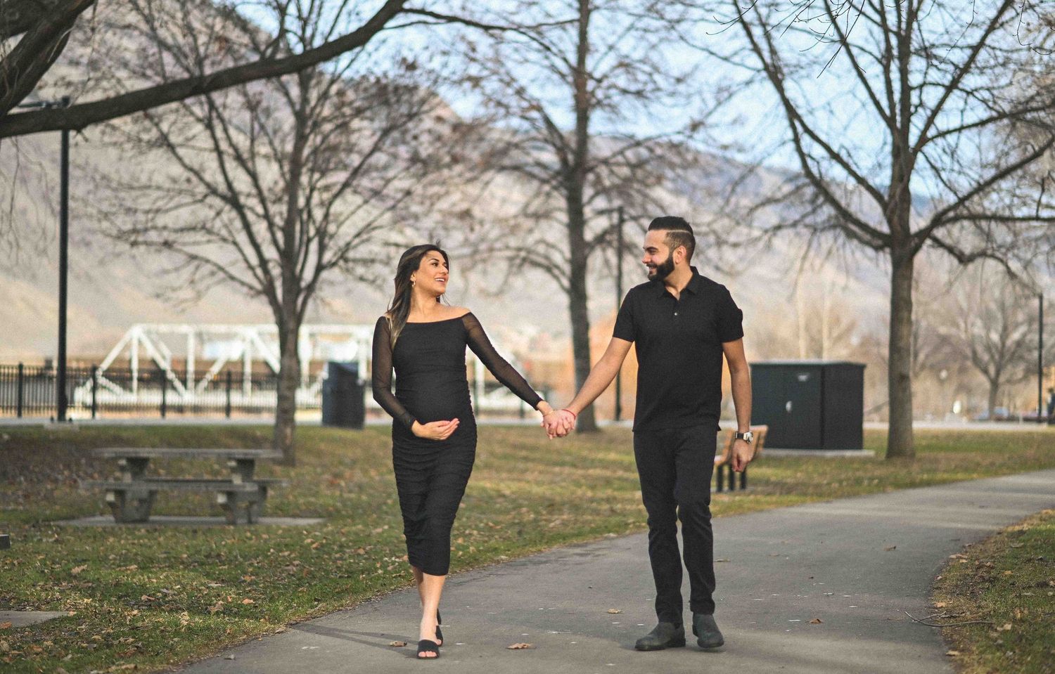 Pregnant couple walking hand in hand through a park in matching black outfits.
