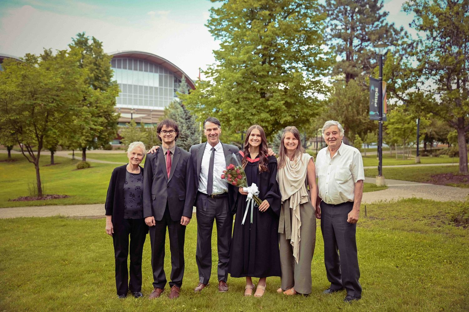 Group of six people posing together outdoors on university campus with graduate in black gown holding flowers.