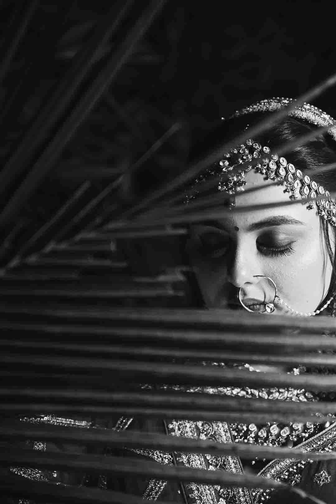 Black and white artistic portrait of Indian bride in traditional jewelry and nose ring behind diagonal lines.