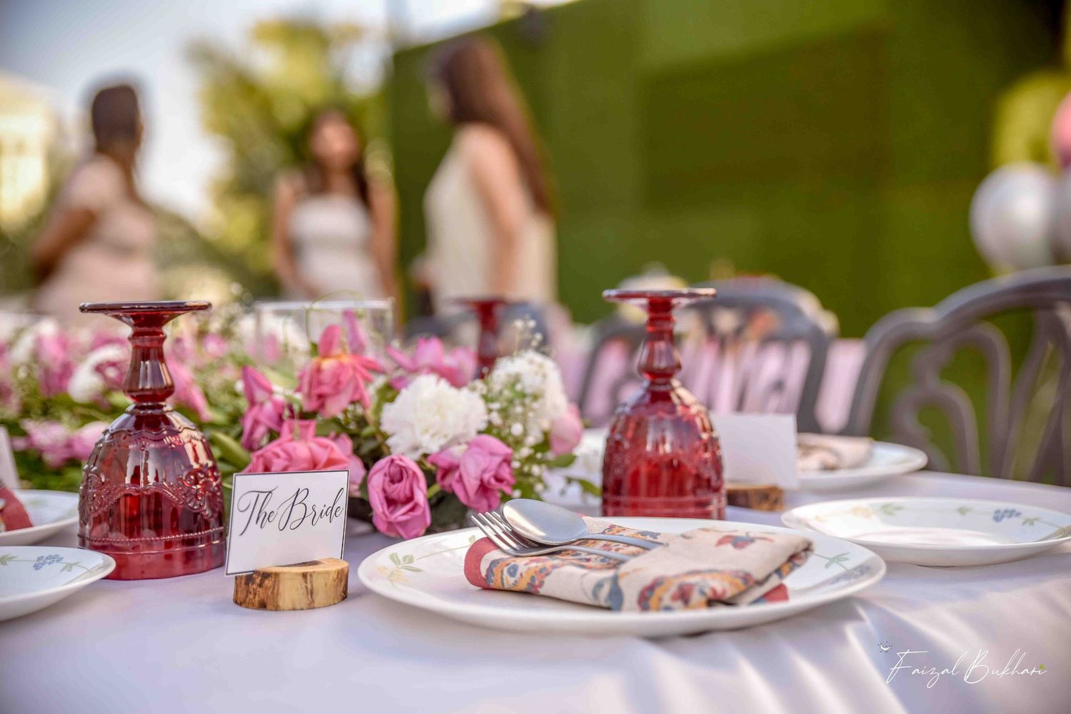 Elegant bridal brunch table setting with pink floral centerpiece, ruby red glassware, and place cards outdoors.