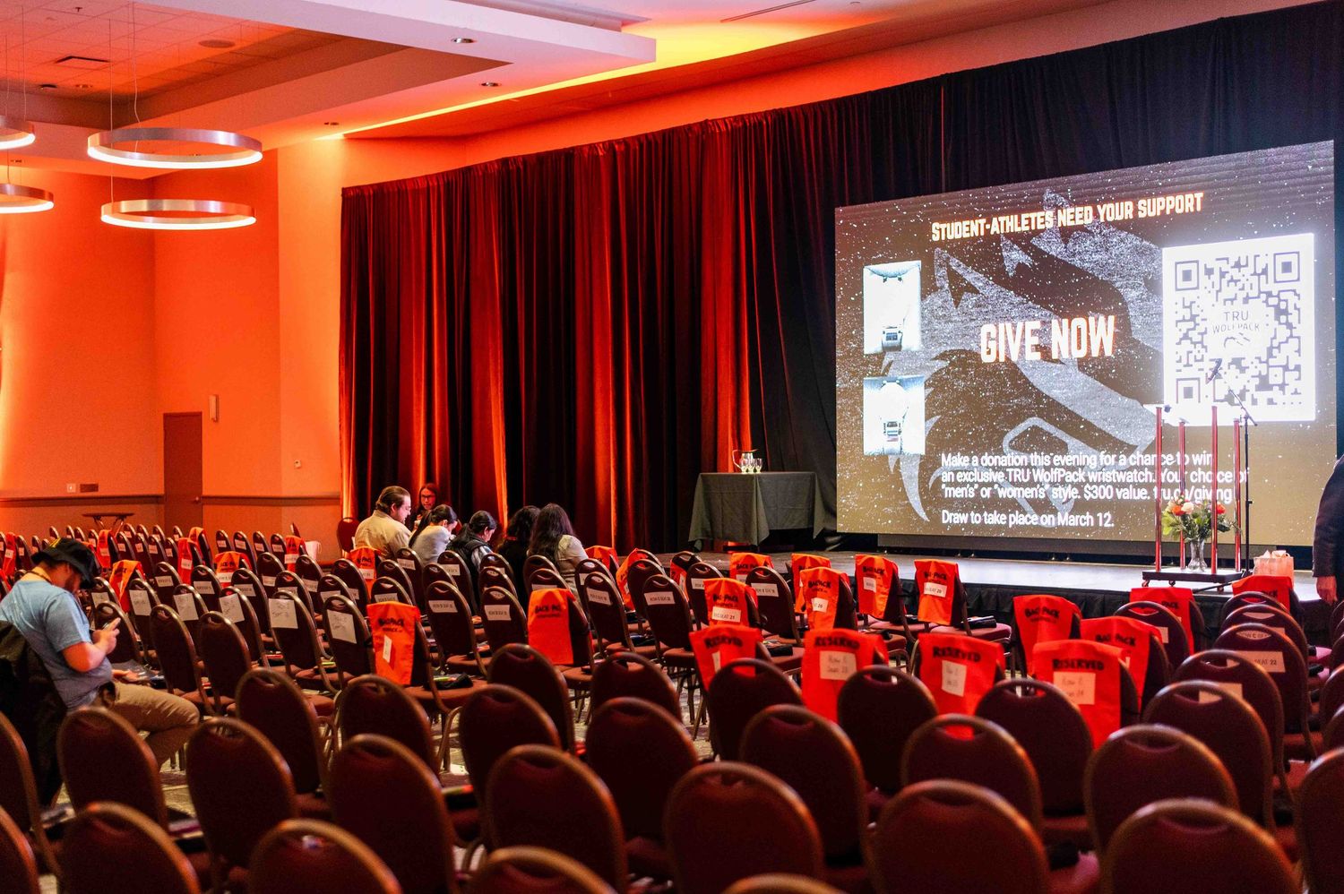 Large conference hall with red seating and a projected screen displaying a student-athlete fundraising presentation.
