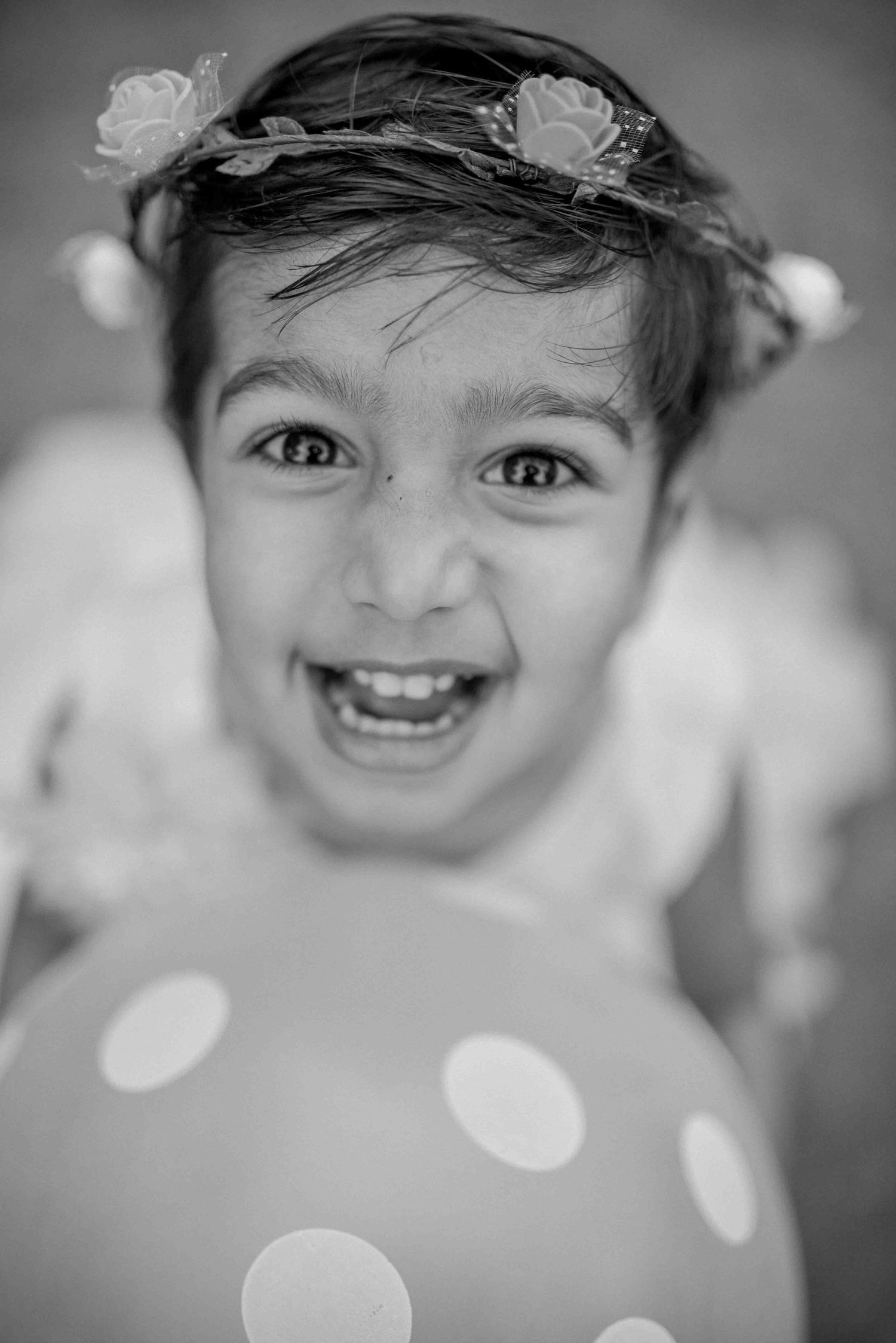 Black and white portrait of a joyful child with floral headband leaning over a polka dot balloon.