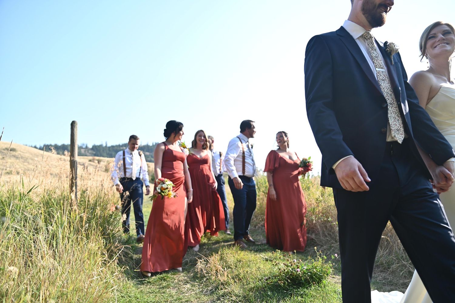 Wedding party walks outdoors in a sunny field, bridesmaids in rust red dresses, groomsmen in navy suits.