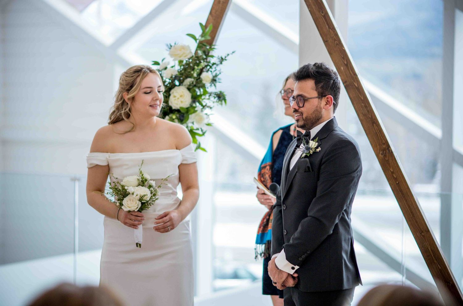 Bride in off-shoulder white gown holds bouquet beside groom in dark suit during indoor mountain wedding ceremony.