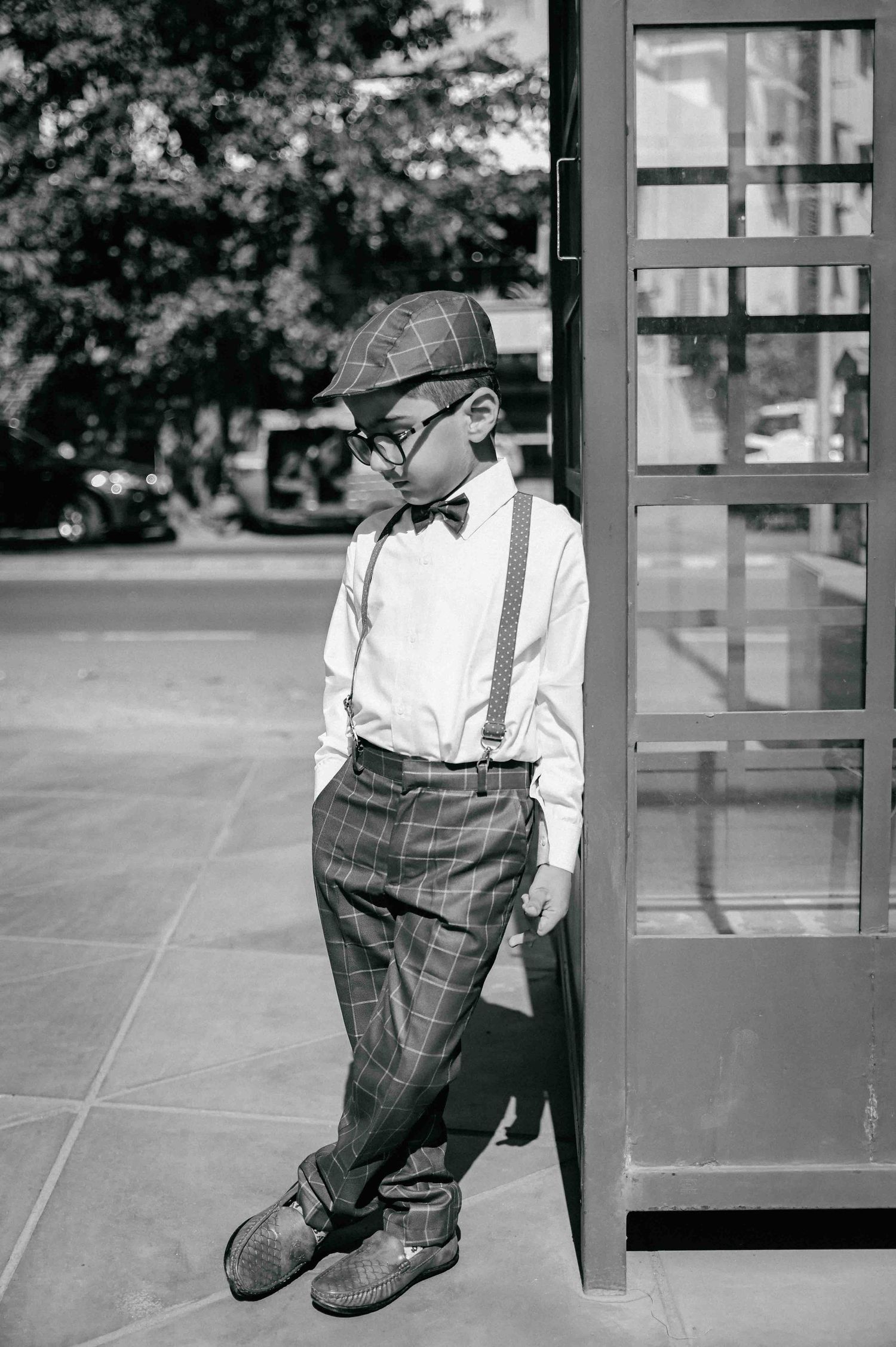 A young boy in vintage plaid outfit with suspenders, bow tie, and cap leans against a door outside.
