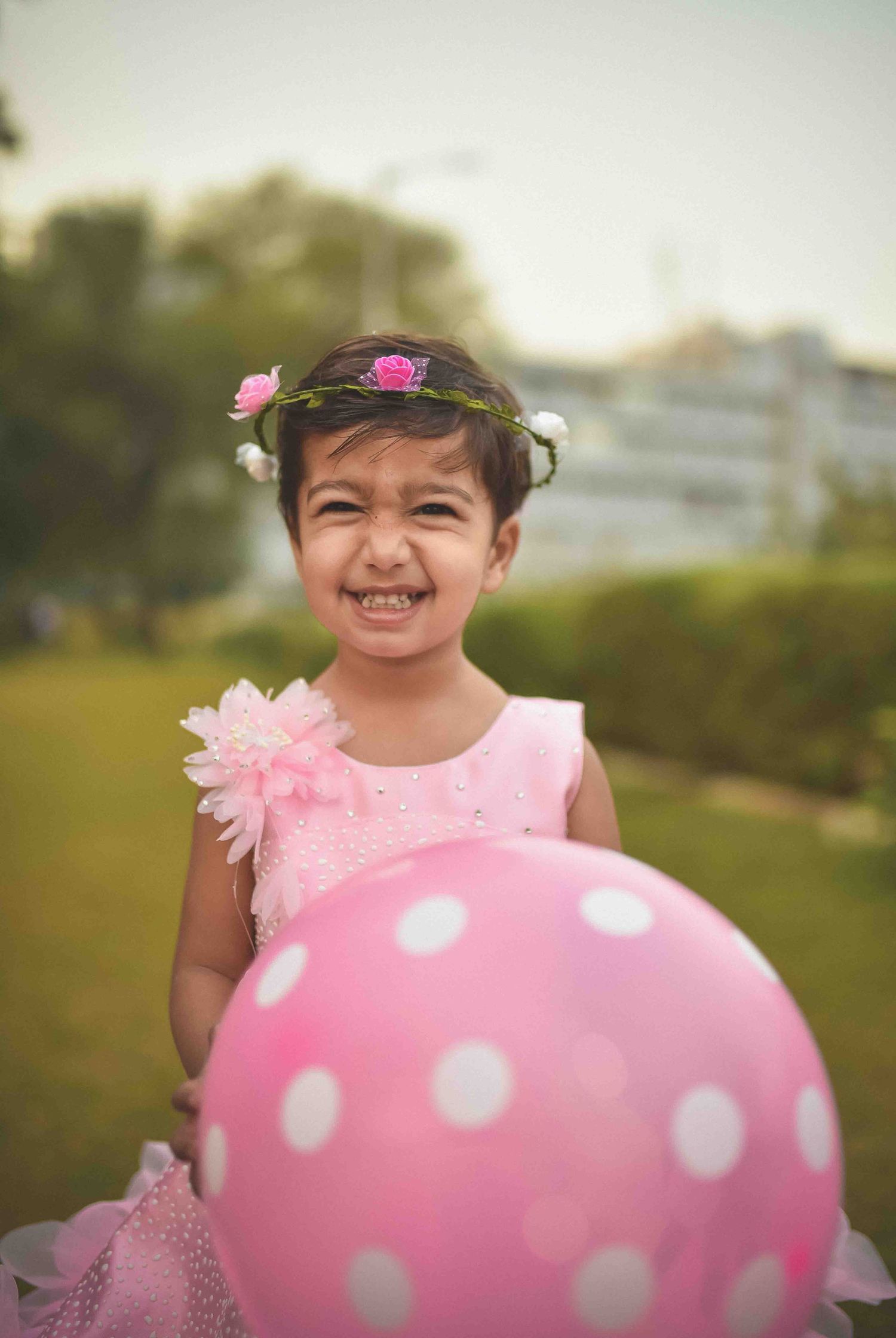 Smiling girl in pink dress wearing floral crown holding large pink polka dot balloon outside.