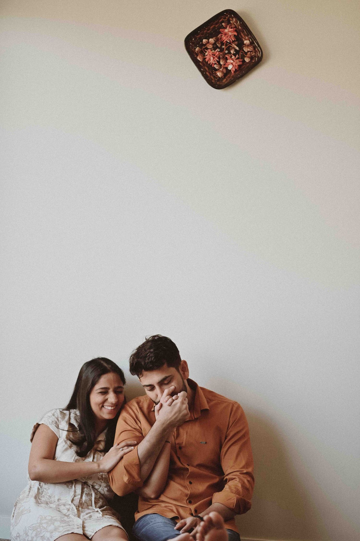 A smiling couple sits together laughing against a white wall with decorative plate above.