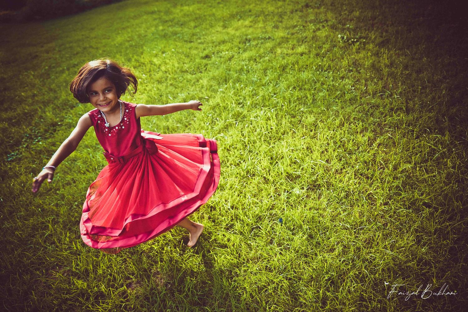 A cheerful young girl in a flowing red dress runs and spins on a bright green grassy field.