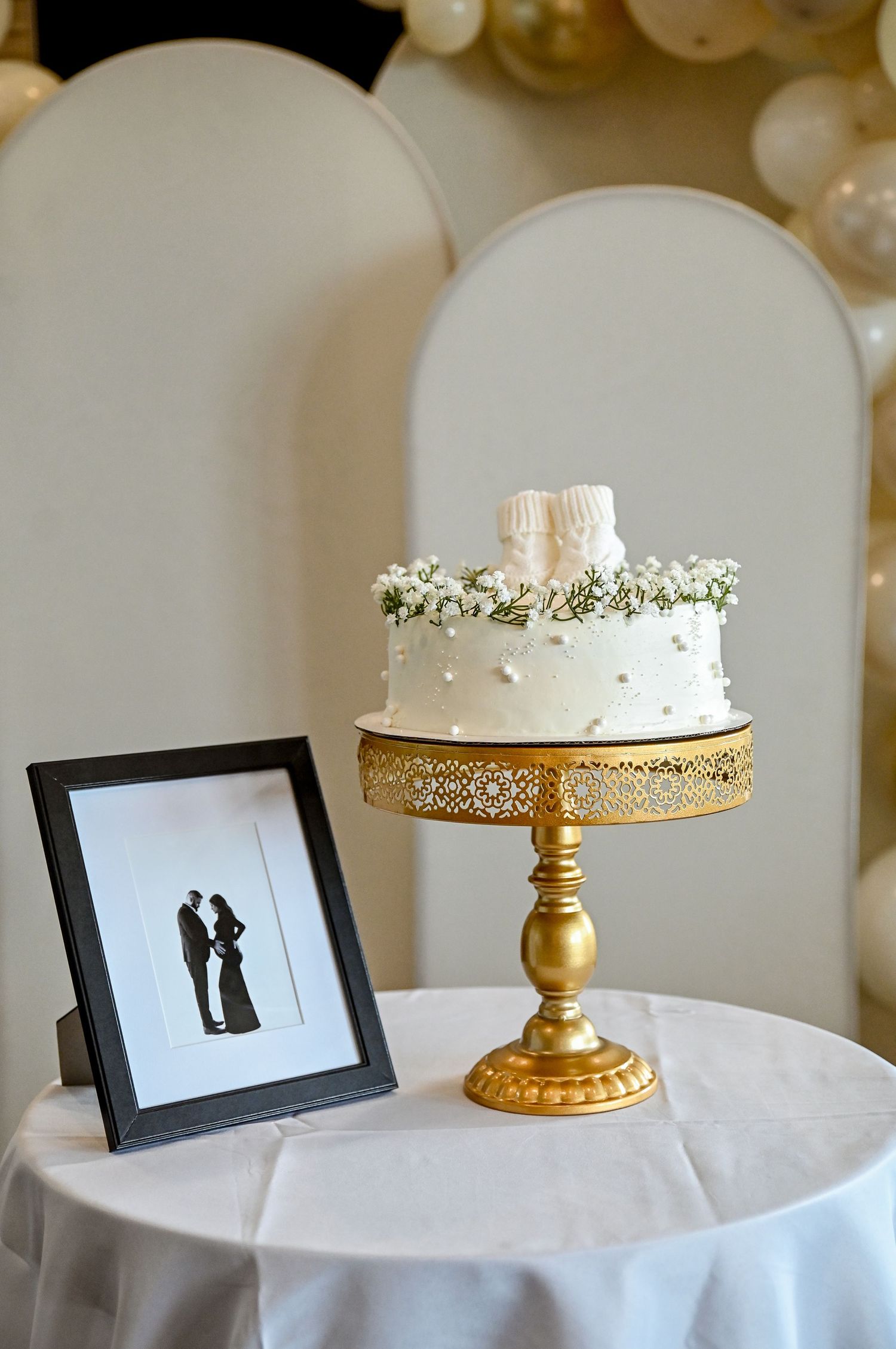 White baby shower cake on gold stand with tiny booties topper beside framed silhouette print on draped table. Kamloops Event Photographer