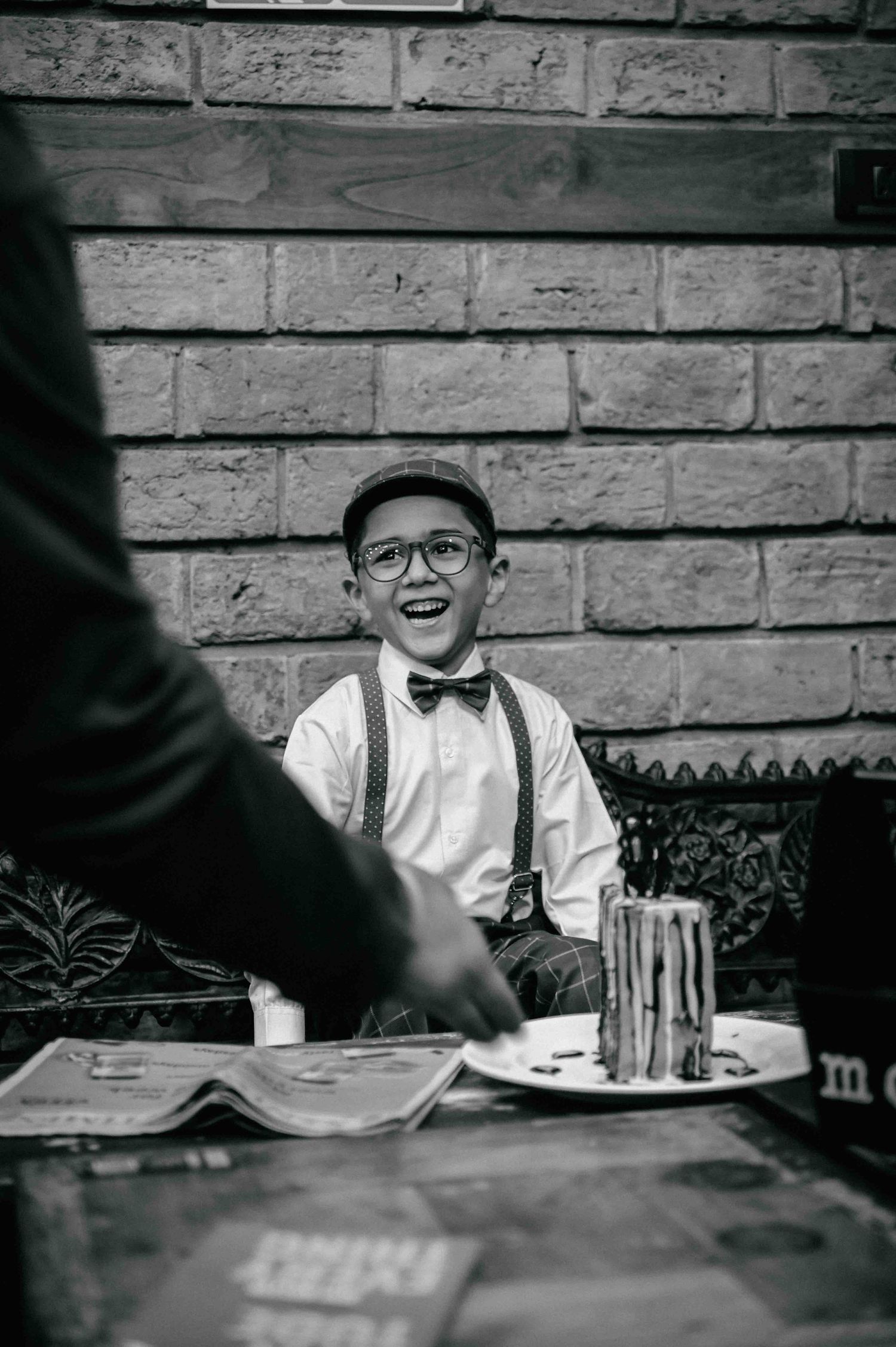 A cheerful boy in vintage suspenders and bow tie laughs at a table with a birthday cake nearby.