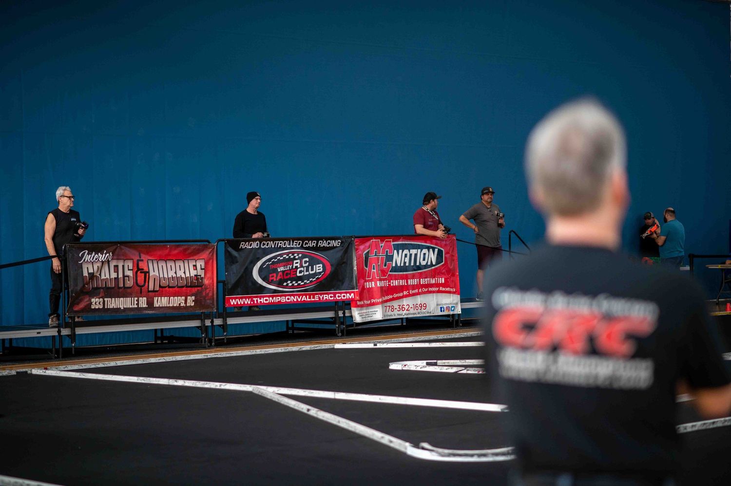 RC racing event spectators watching from behind sponsor banners on an indoor track.