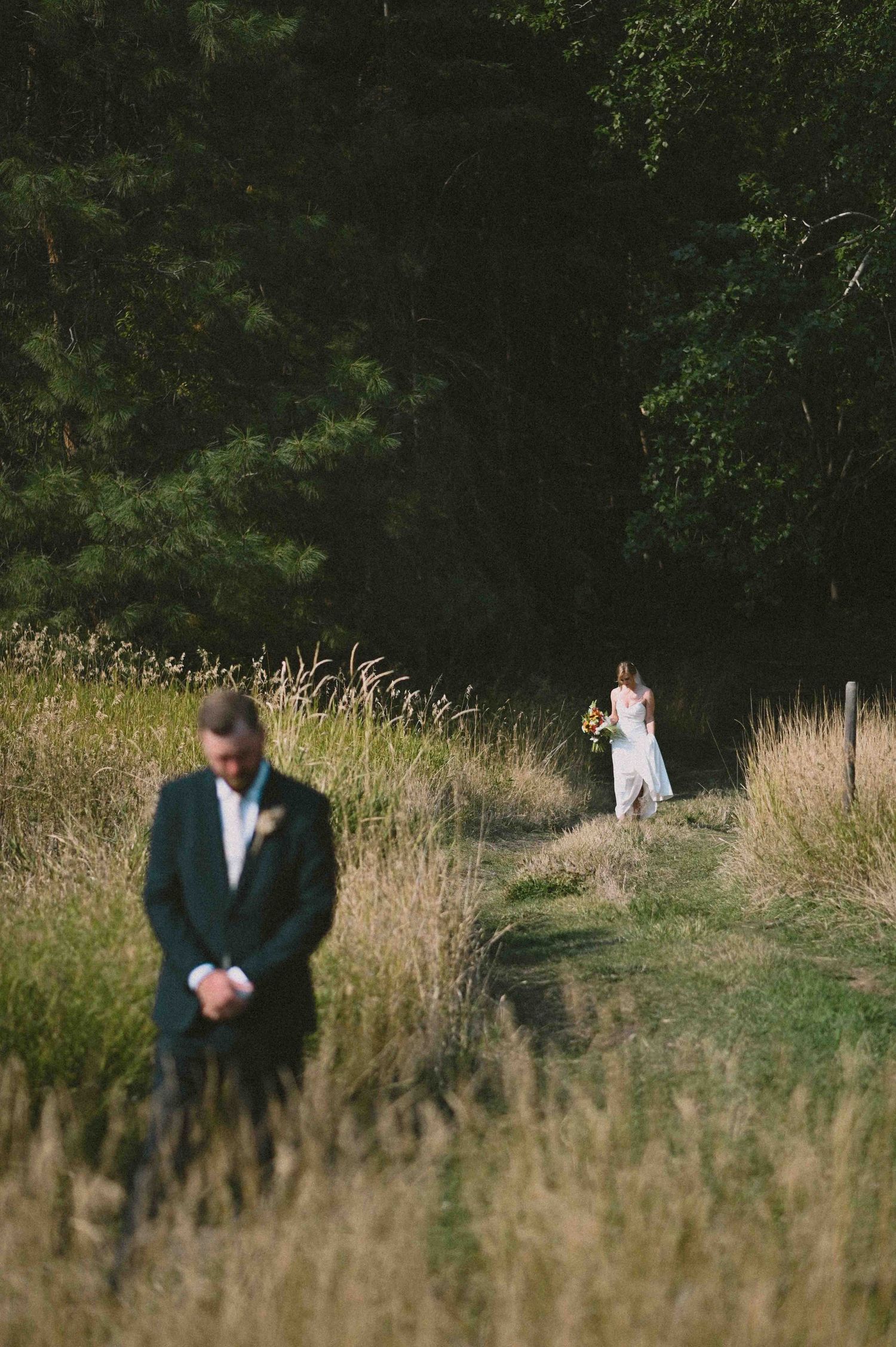 Artistic first look photo of groom in foreground and bride walking toward him through tall golden grass.