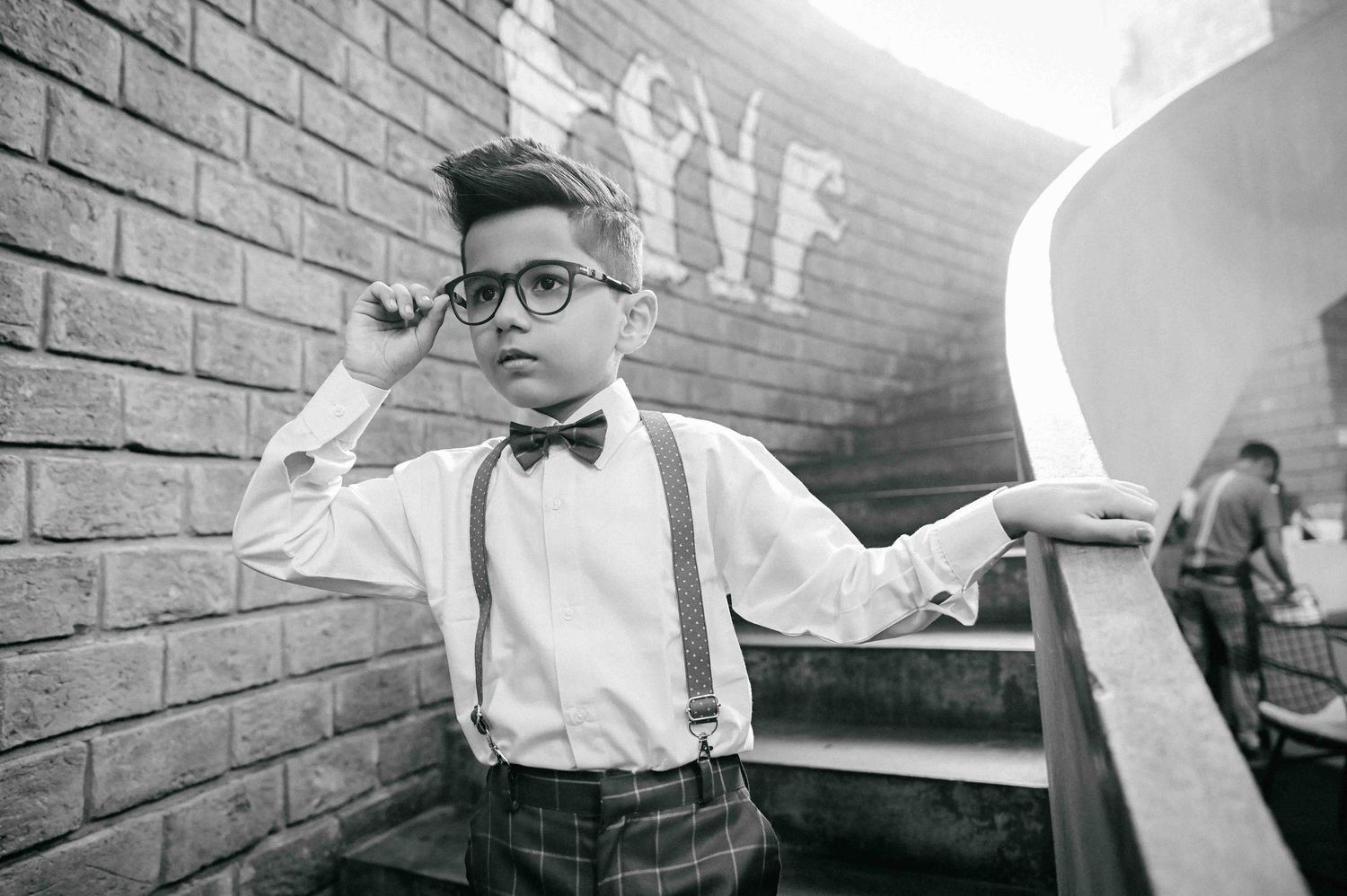 Stylish boy in glasses, bow tie and suspenders posing on brick staircase in black and white.