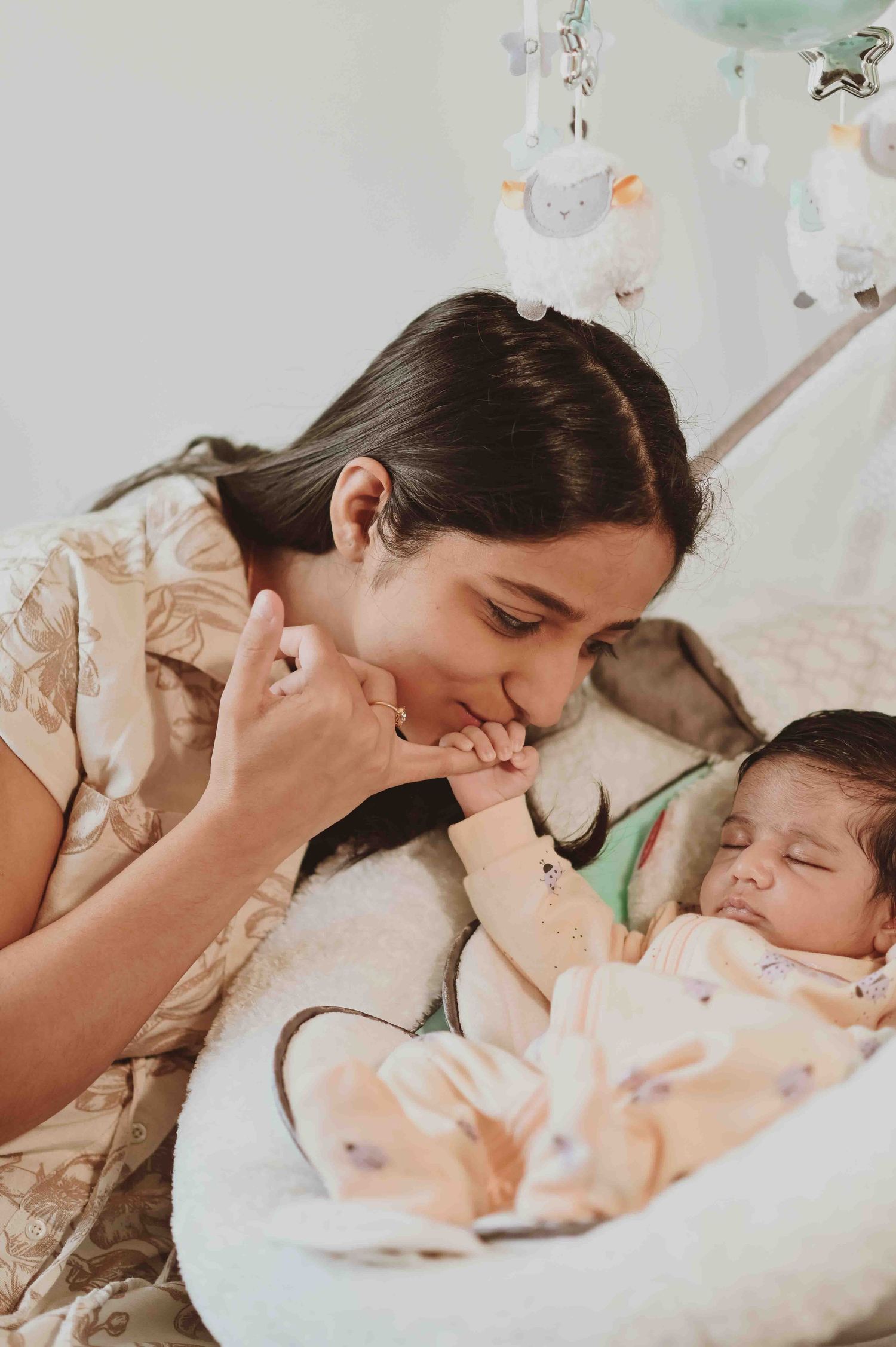 A mother lovingly kisses her sleeping newborn baby's hand in a baby swing.