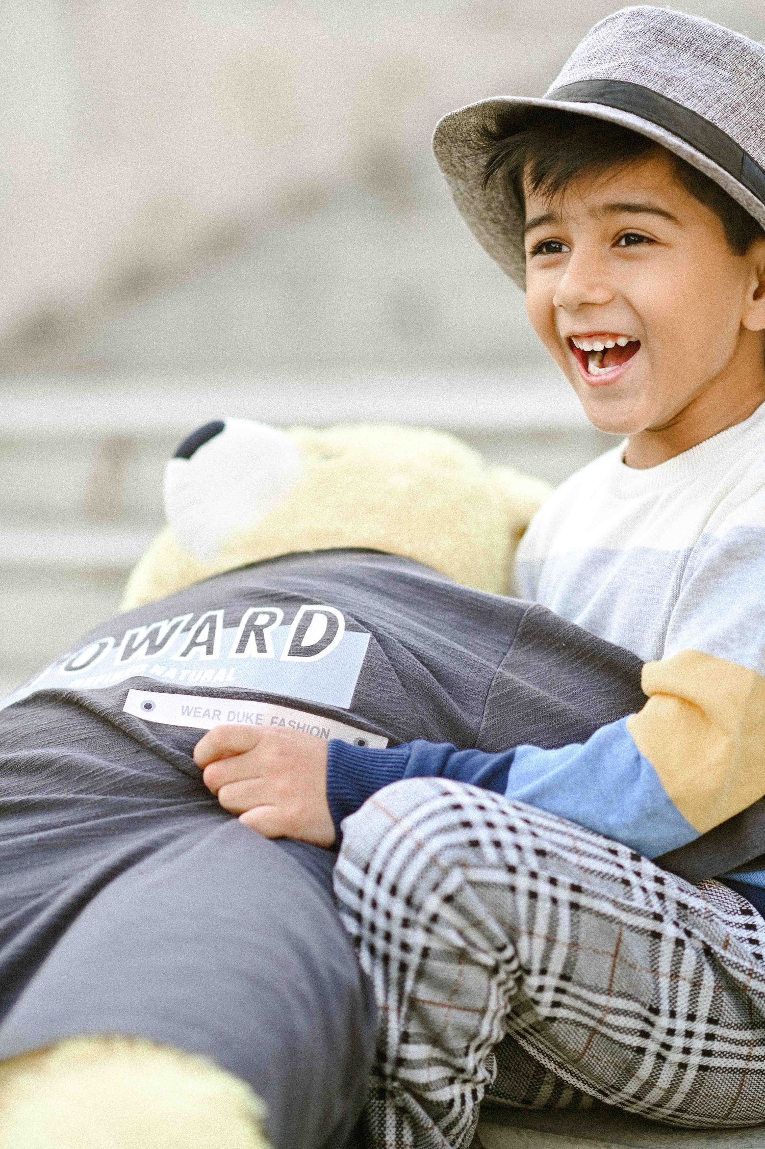 Joyful boy in fedora hat laughing while holding large teddy bear on outdoor steps.