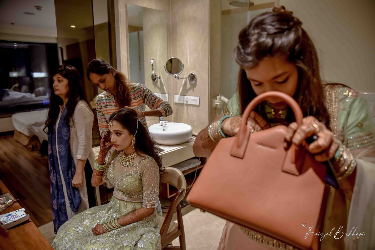 Indian bride in embroidered lehenga holding a tan handbag while bridesmaids assist in hotel getting-ready room.