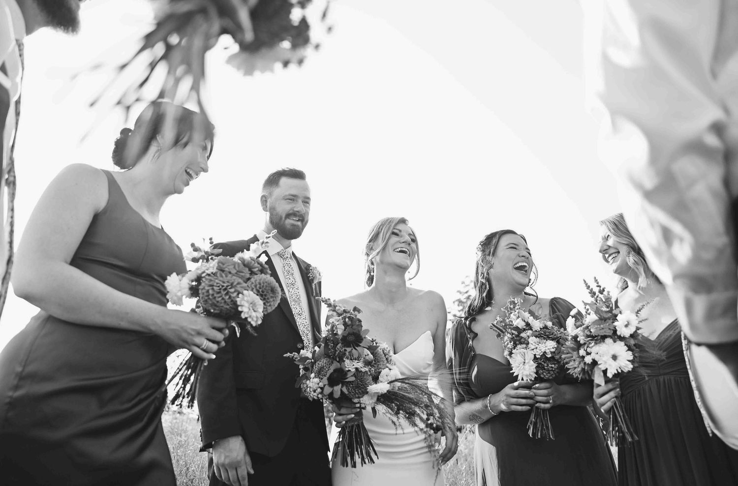 A joyful bride and groom laugh with their wedding party in a candid black and white celebration photo.