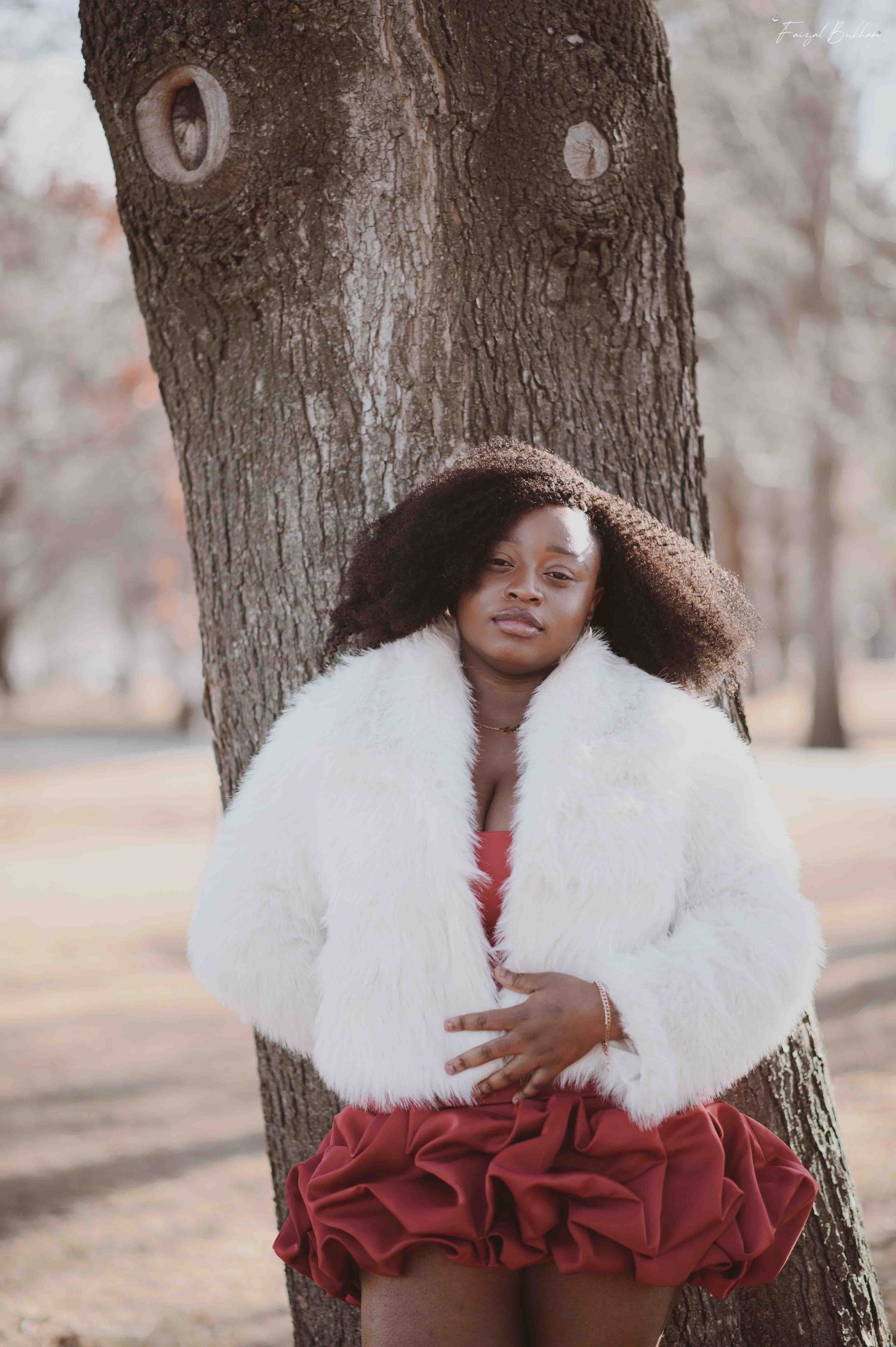 Confident Black woman in white fur coat and red ruffled skirt leaning against a tree in park.