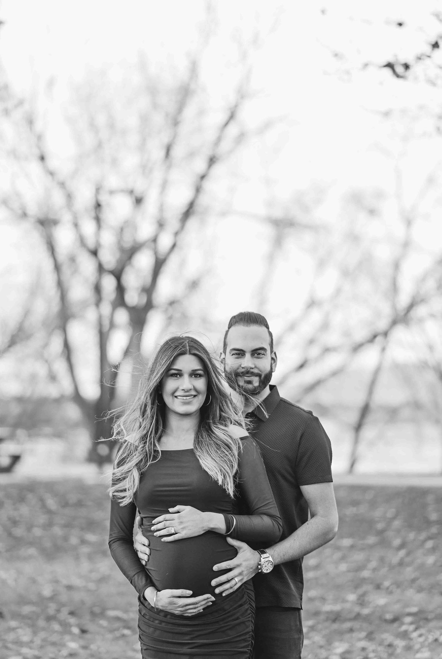 Black and white portrait of smiling couple cradling pregnant belly in park.