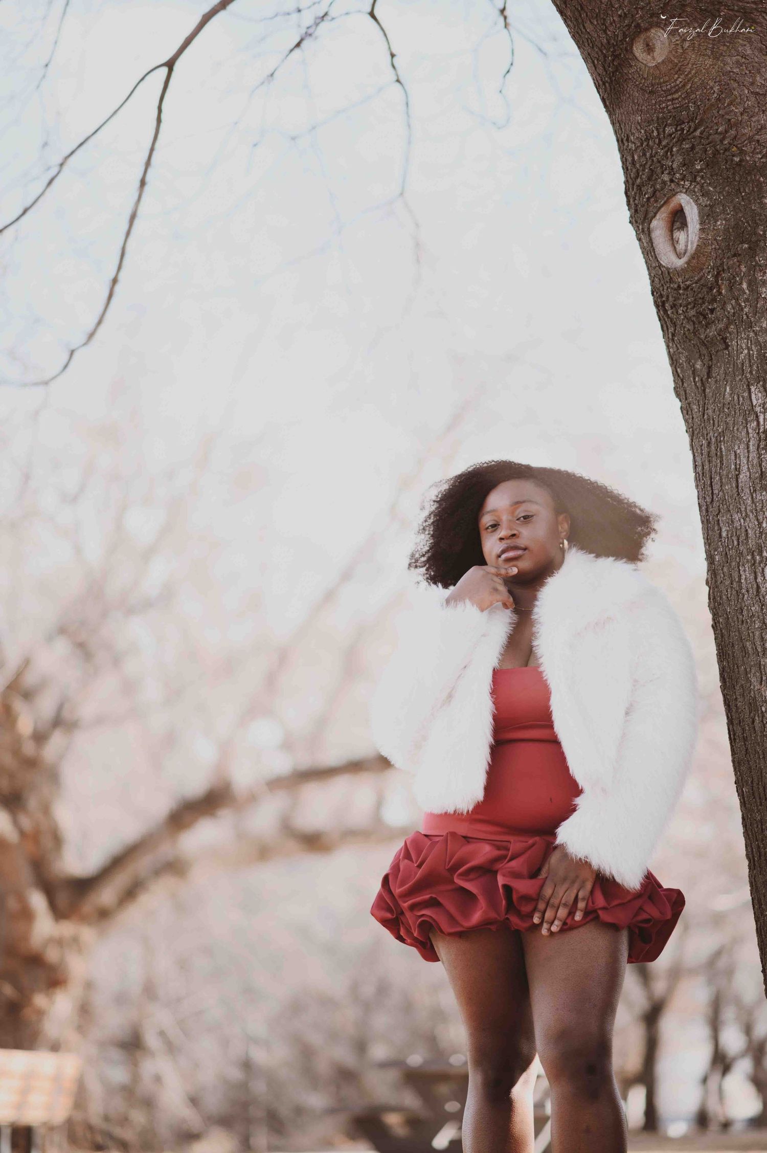 Young Black woman in white faux fur jacket and red ruffled skirt posing gracefully beside a tree.