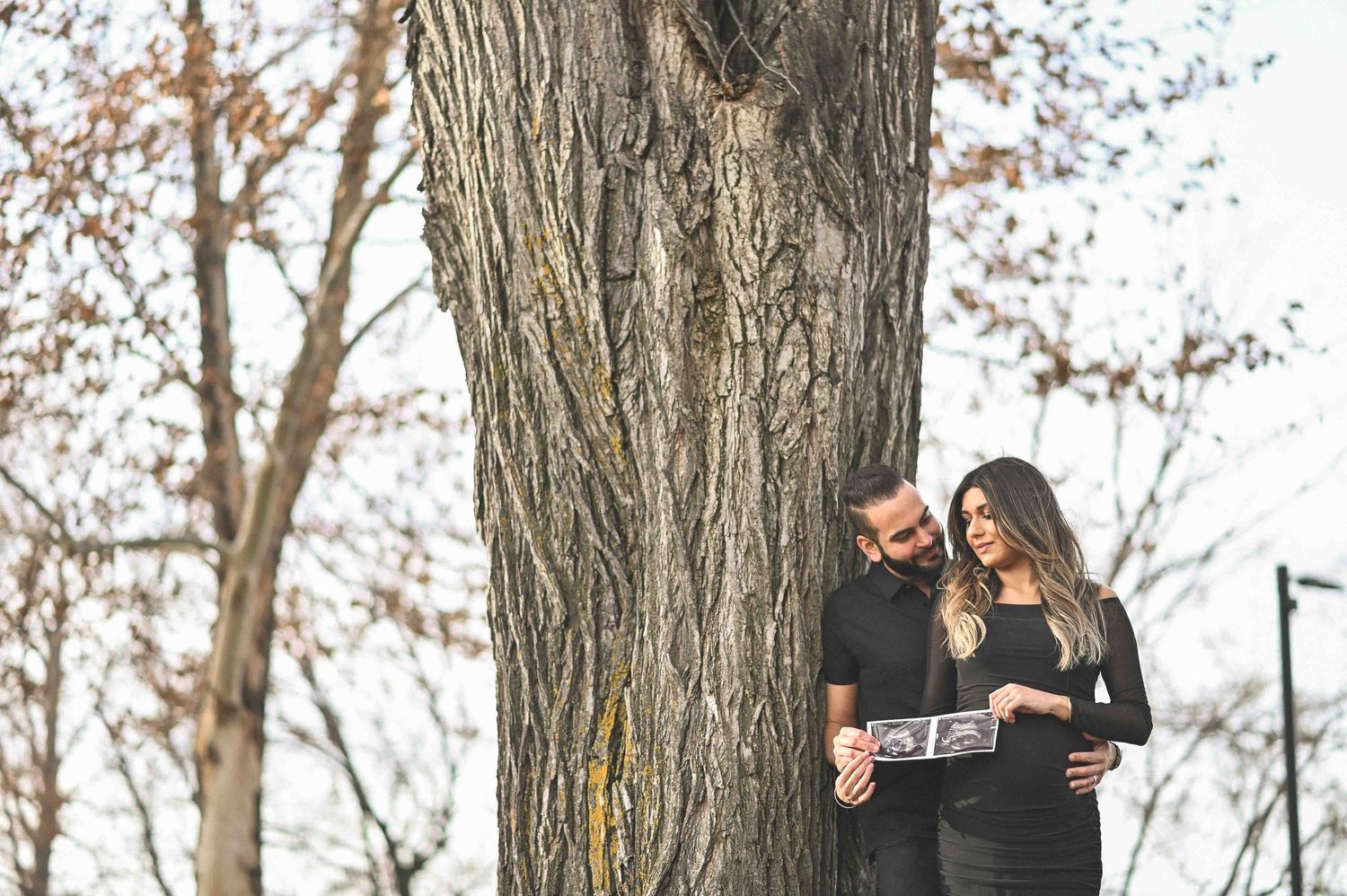 Expecting couple leaning against a tree holding ultrasound photos in a park.