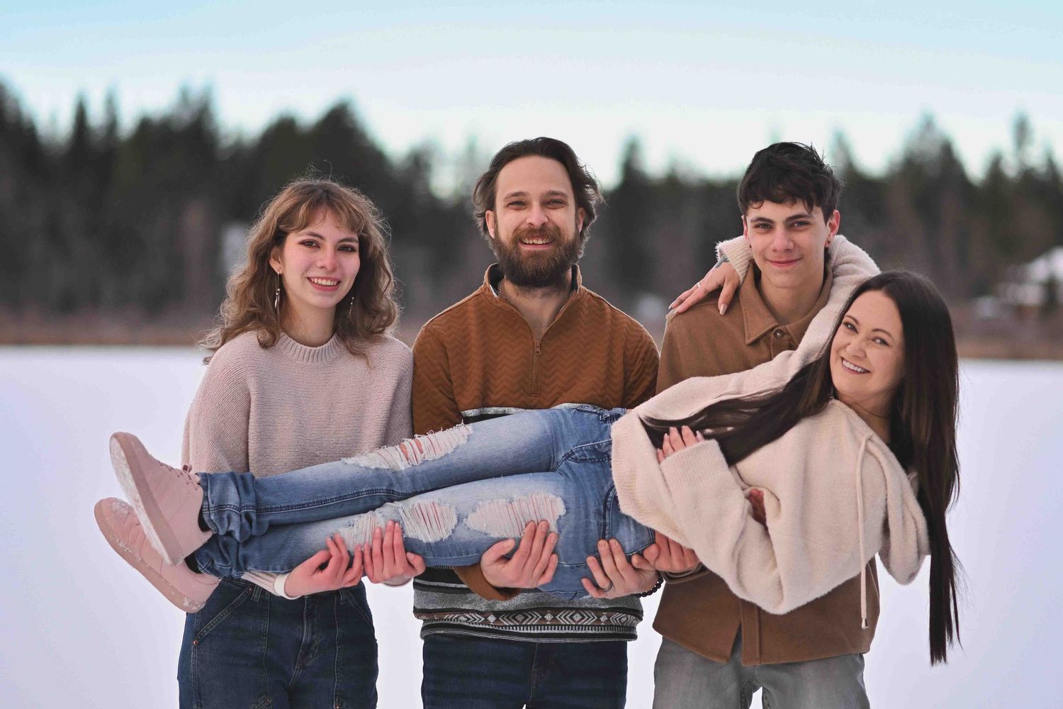 Group of four friends posing playfully in winter snow, with two people carrying a woman horizontally outdoors.