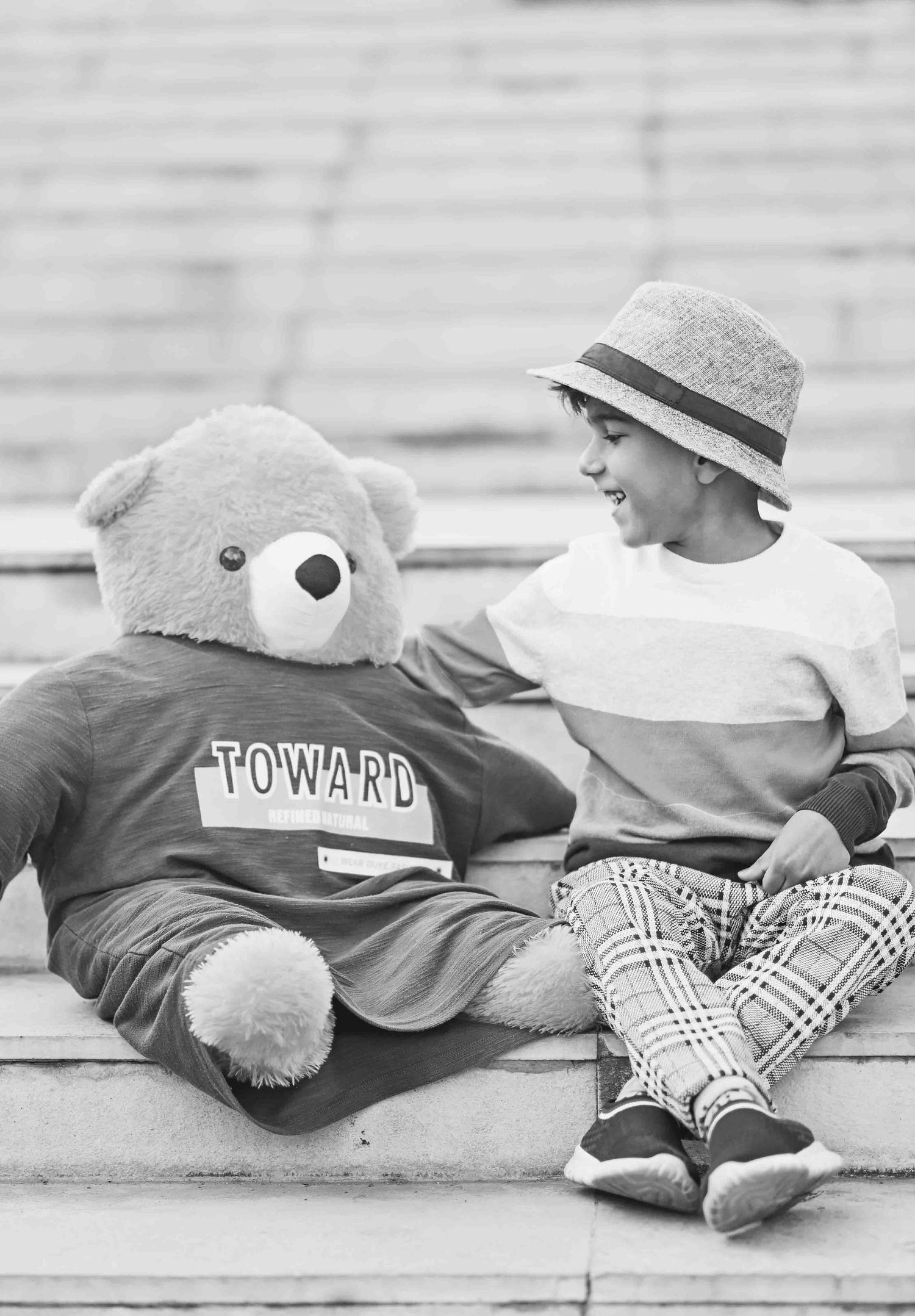 Smiling boy in casual striped shirt and plaid pants sitting on steps beside large teddy bear.
