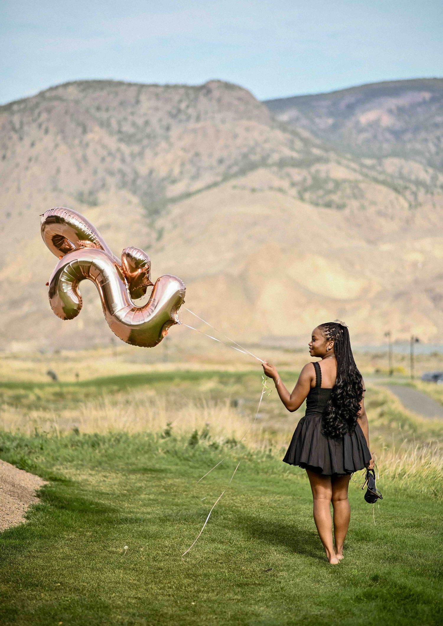 Young woman in black dress holding rose gold number balloons on green hillside with mountains.