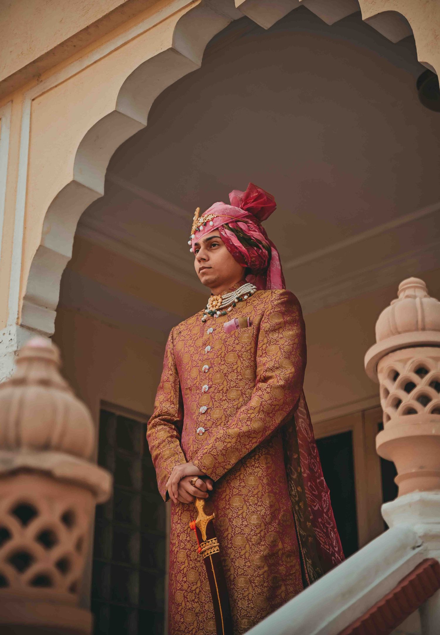 An elegant Indian groom in ornate golden sherwani and red turban stands regally under an arched doorway at a palace.