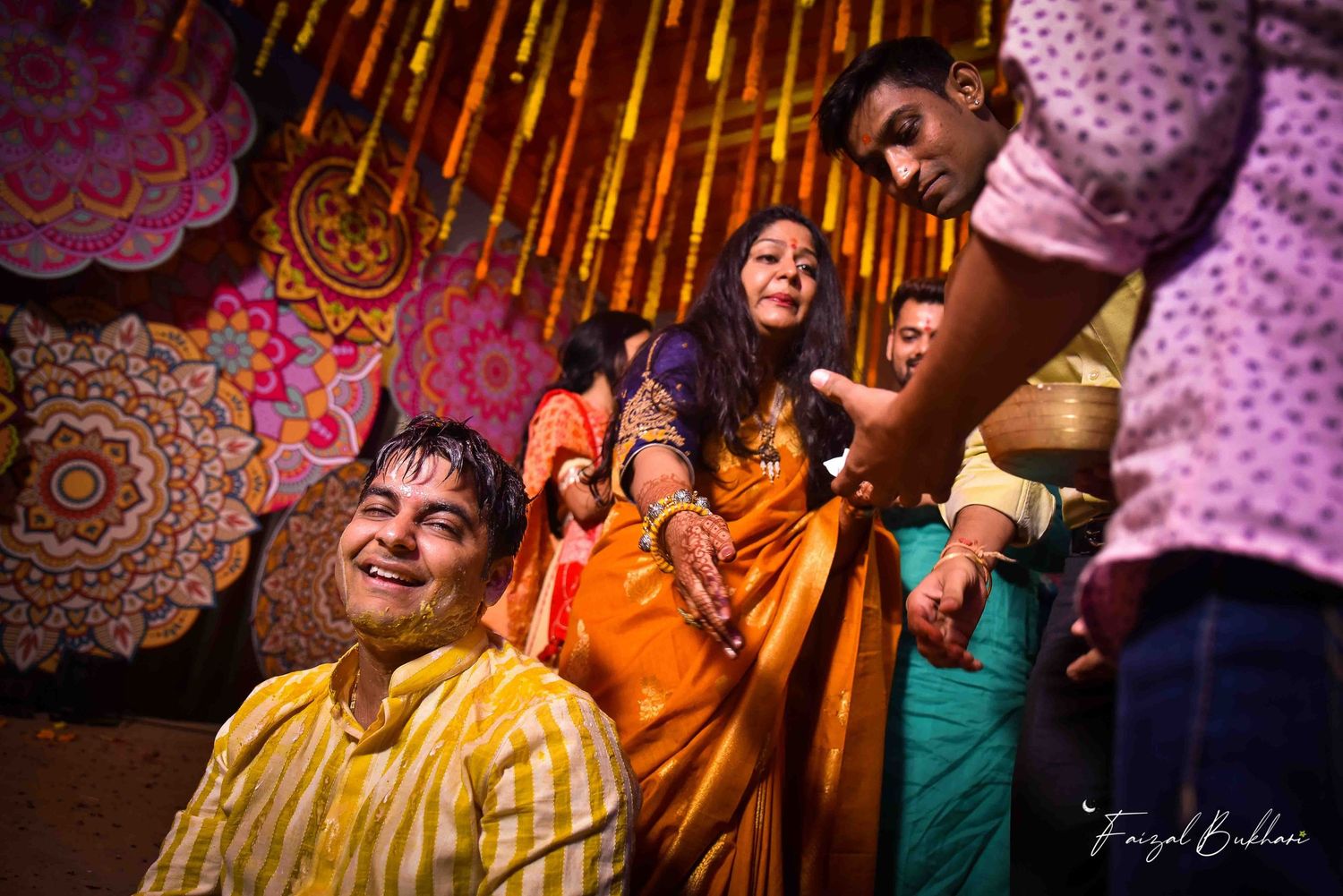Indian groom in yellow outfit laughing during haldi ceremony with family members around him.