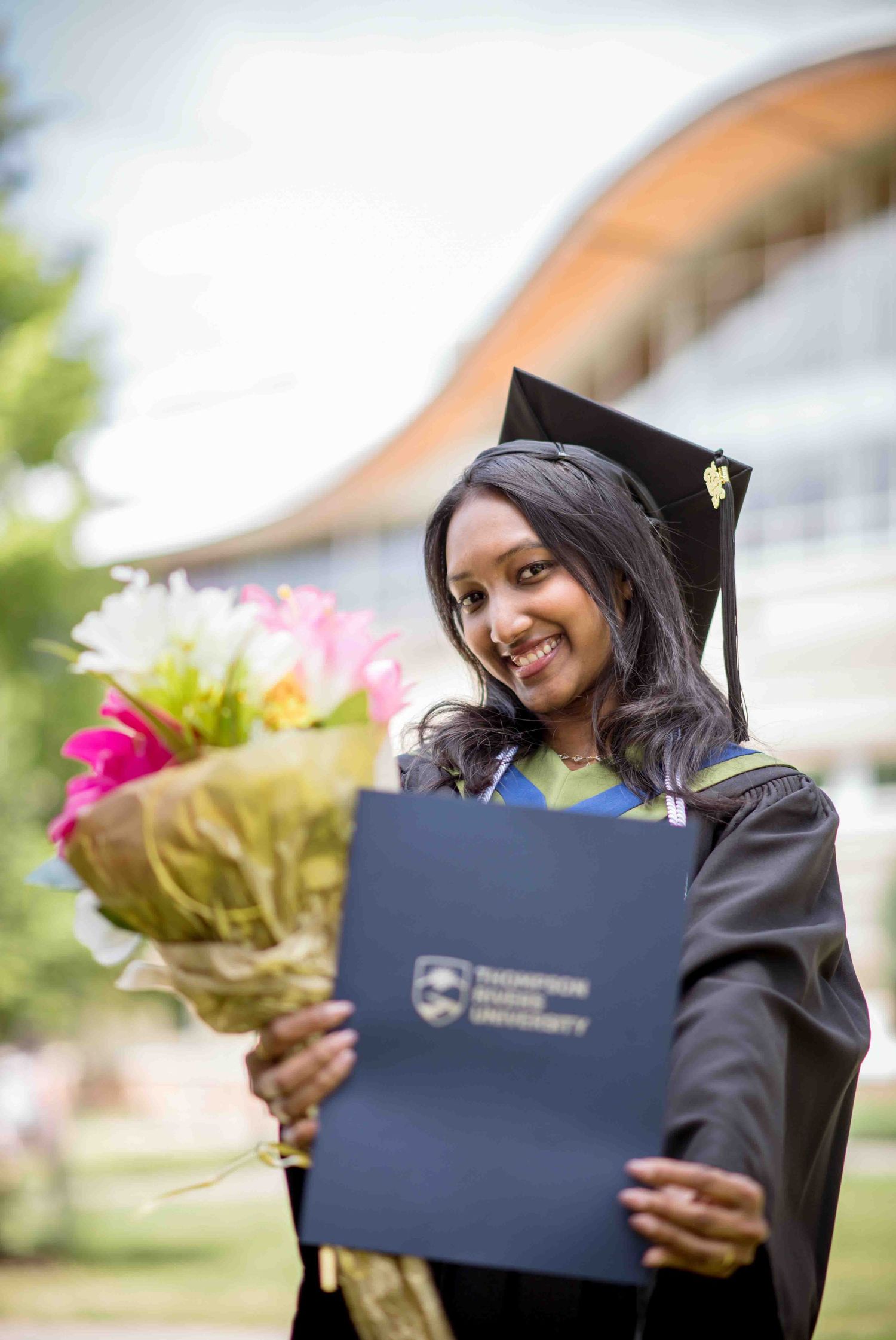 Smiling graduate in cap and gown holds flowers and Thompson Rivers University diploma outdoors.