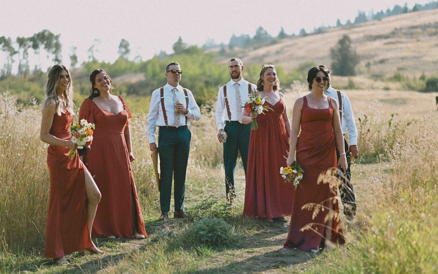 Wedding party in rust-colored dresses and suspenders walks through sunny hillside meadow with bouquets and a dog.