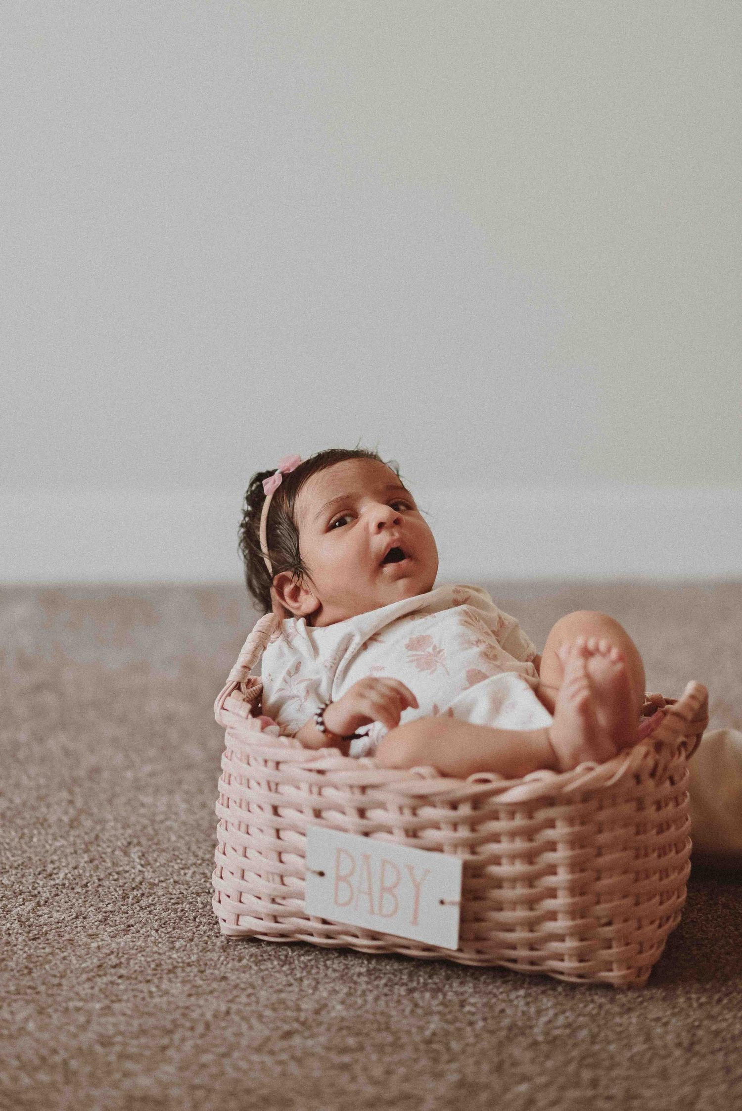Adorable newborn baby sitting in pink wicker basket labeled BABY, wearing floral outfit with hair bow.