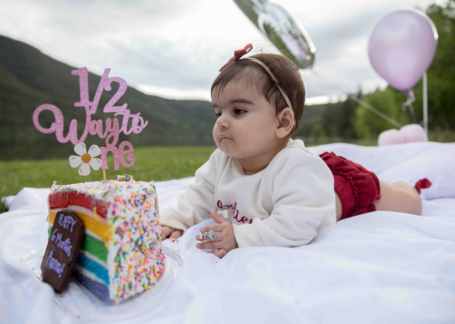 Baby girl lying on white blanket looking at rainbow cake with half way to 1 topper.