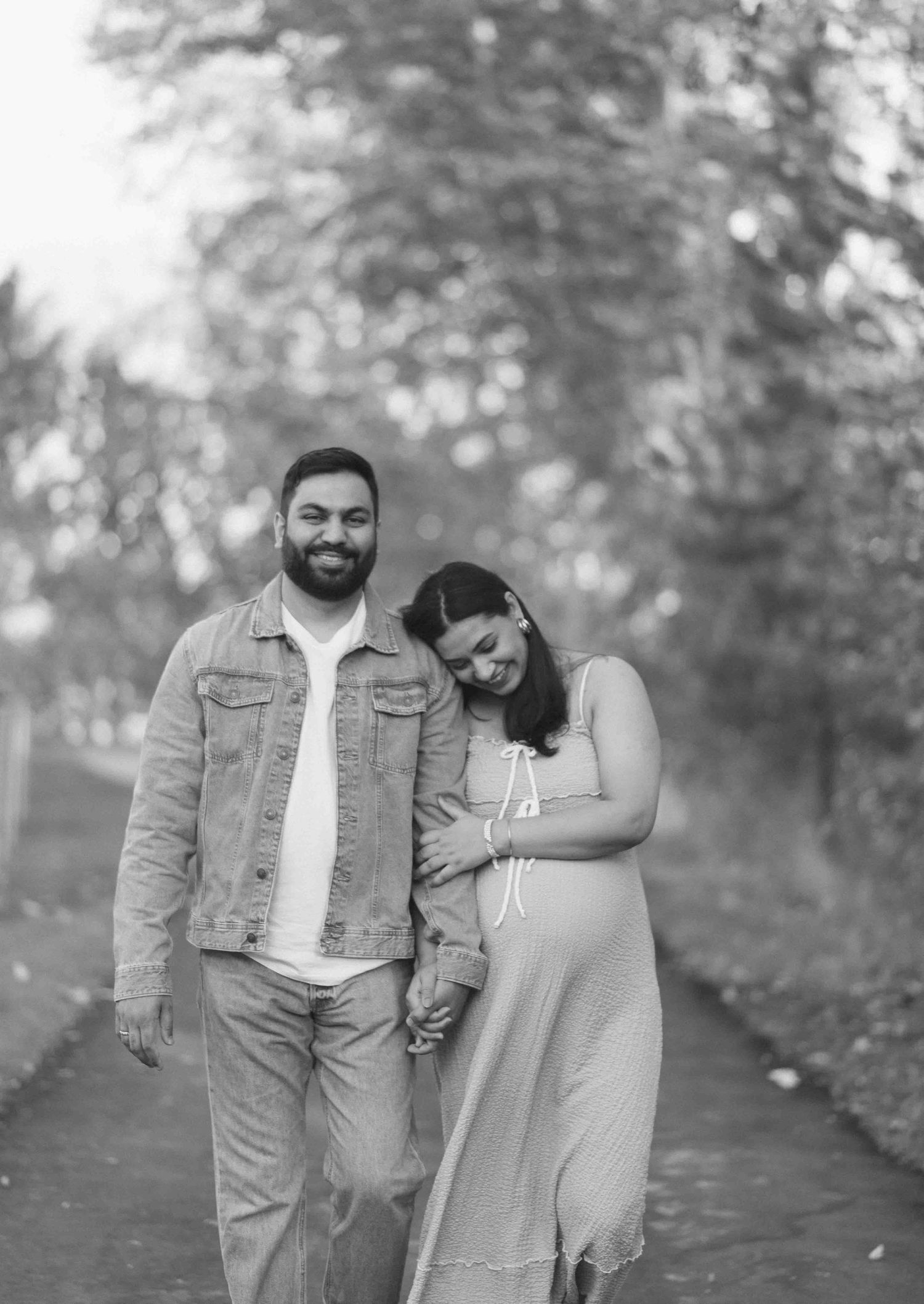Couple walking hand in hand on tree-lined path in black and white maternity photo.
