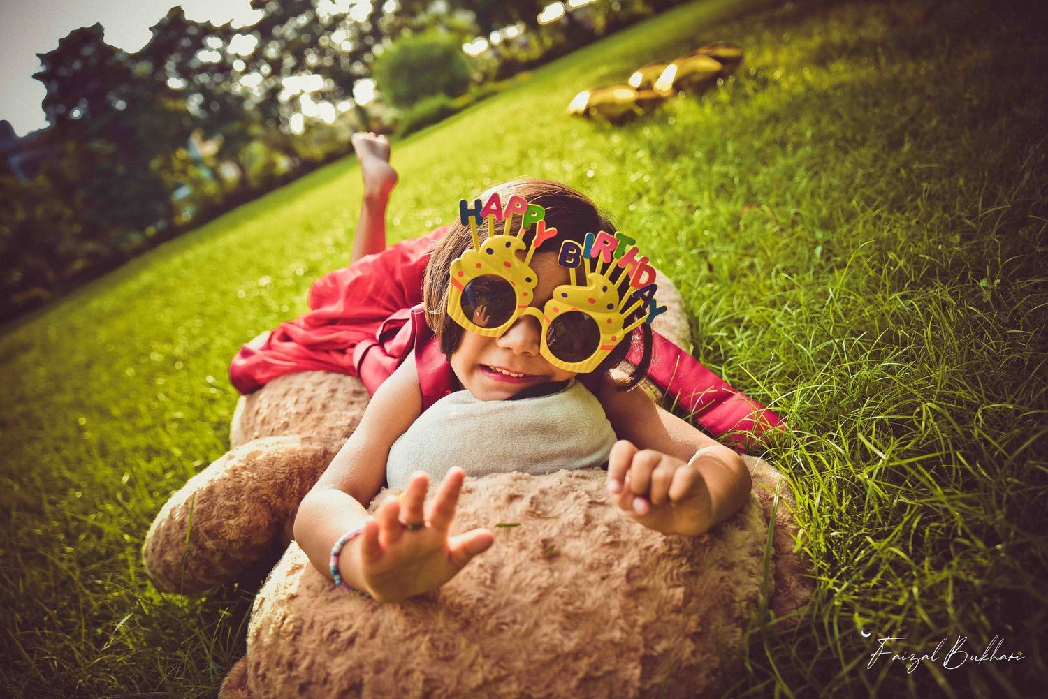 A joyful child lying on a large teddy bear in a green park, wearing festive novelty glasses outdoors.