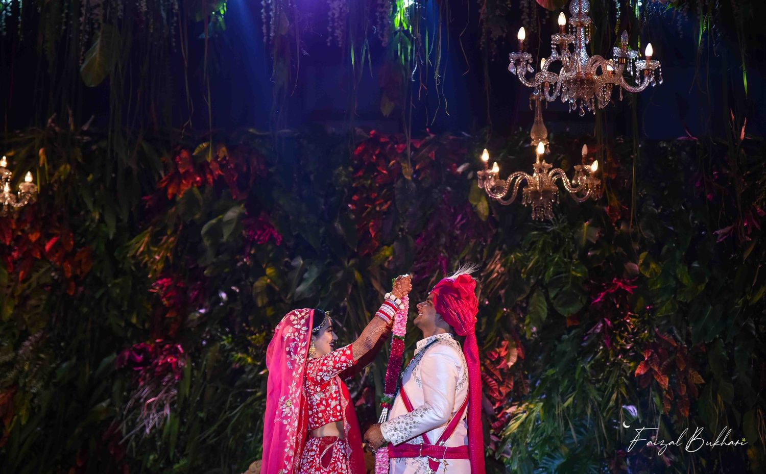 Indian bride and groom sharing a romantic moment under lush floral canopy with glowing chandeliers at night wedding.