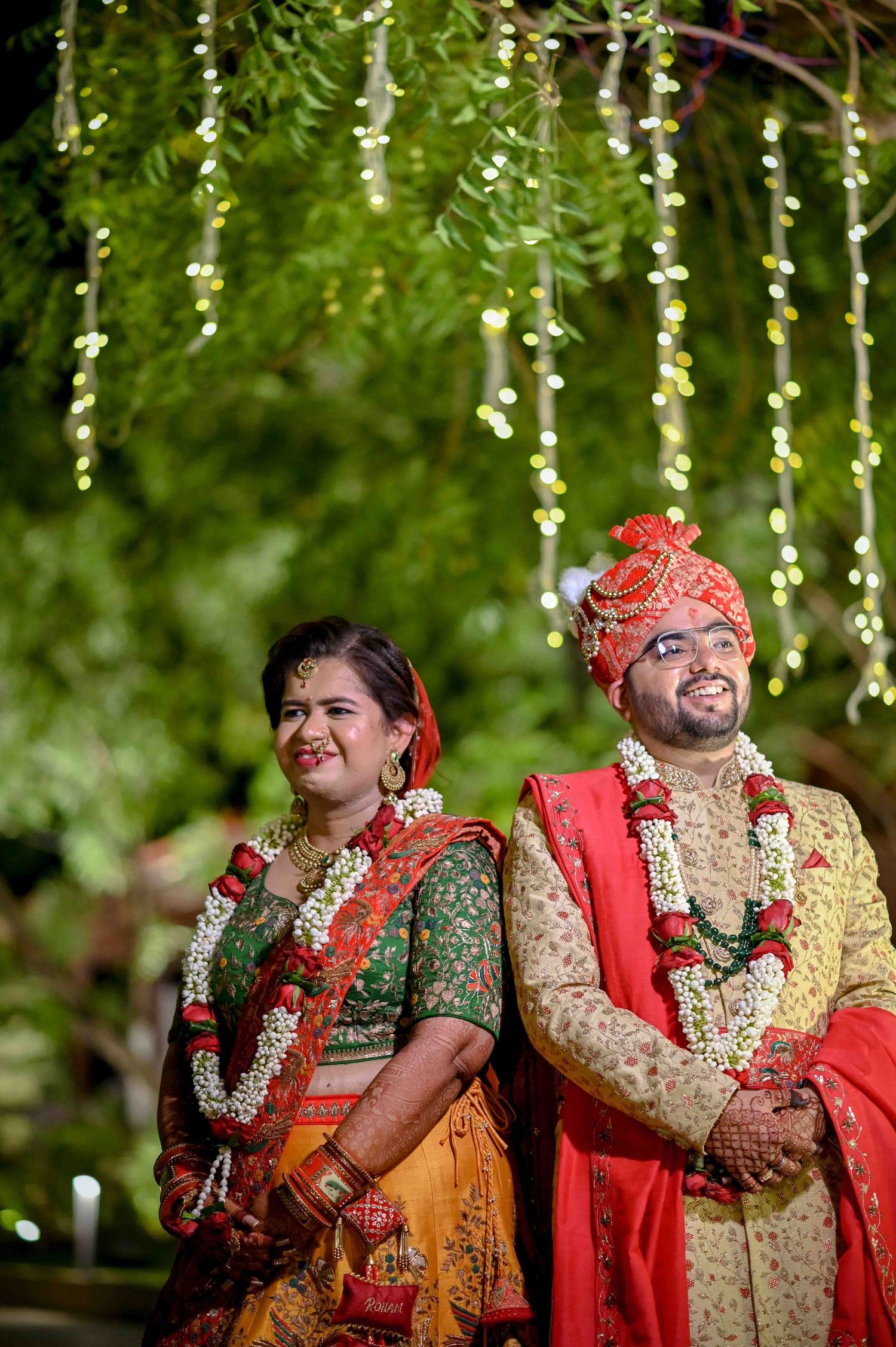 Indian bride and groom in traditional red and gold wedding attire standing under hanging floral fairy lights.