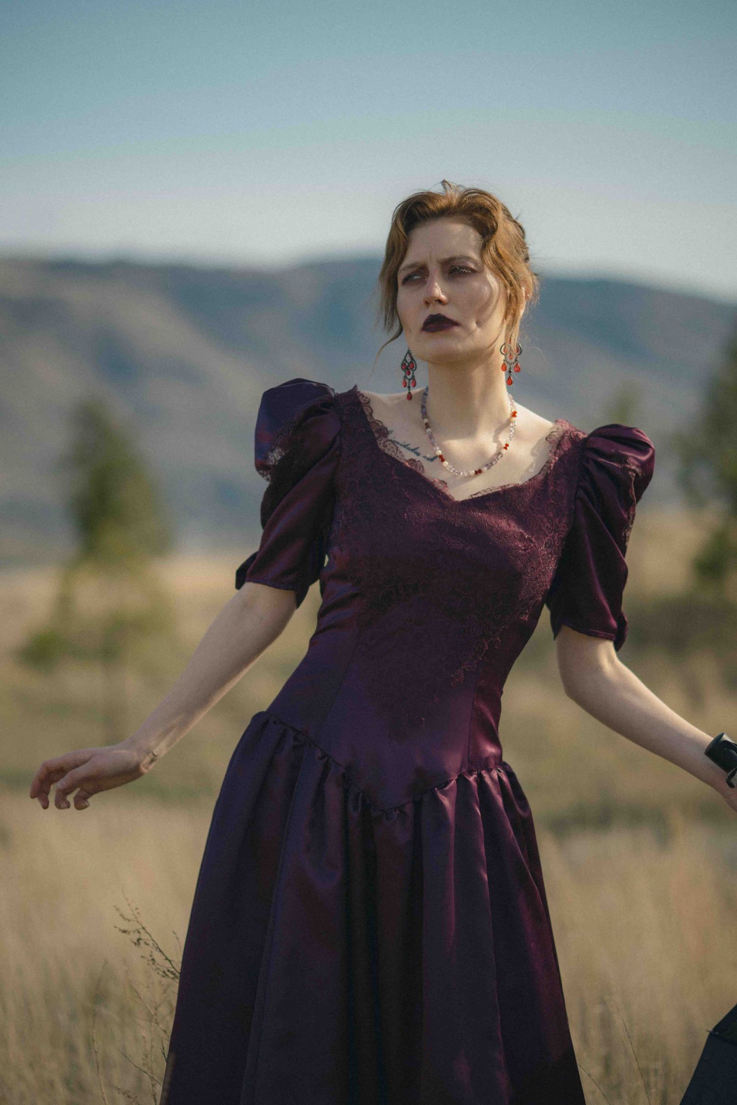 Woman in dark purple Victorian dress with dark lipstick and jeweled earrings poses in dry grassy field.