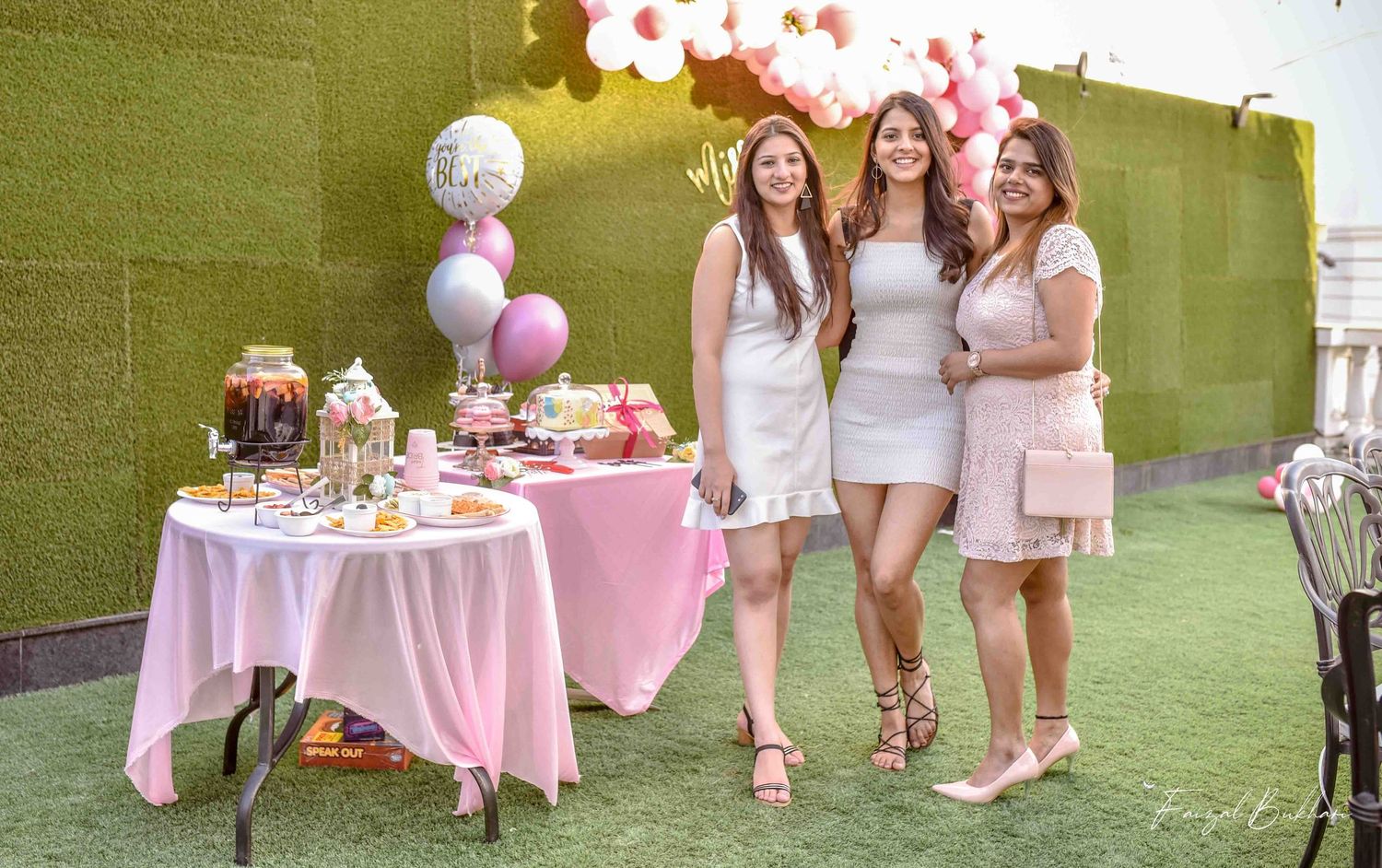 Three women in white and pink outfits smiling at a pink-themed bridal shower dessert table outdoors.