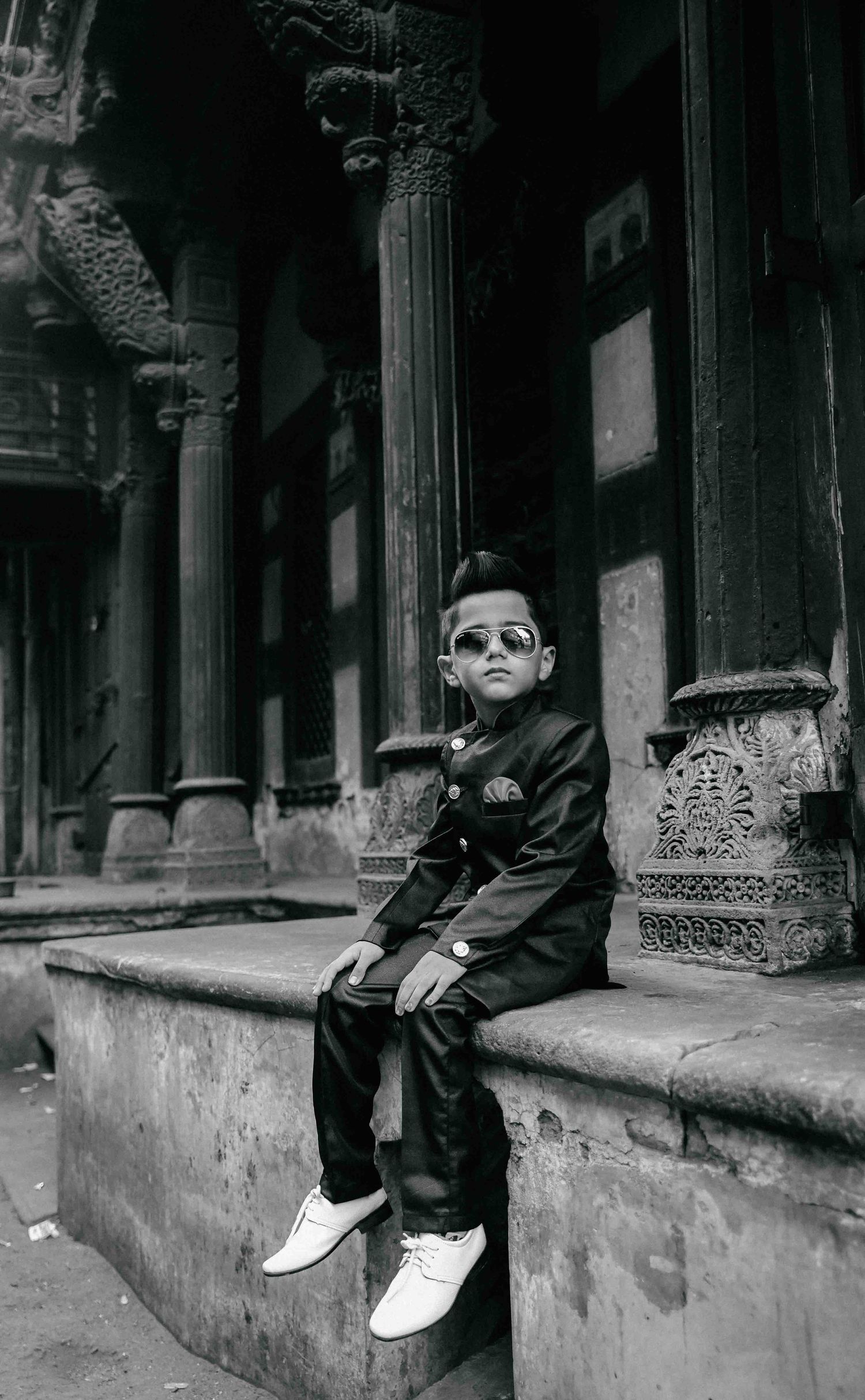 Fashionable young boy in dark suit and sunglasses sitting on a ledge before historic carved columns.