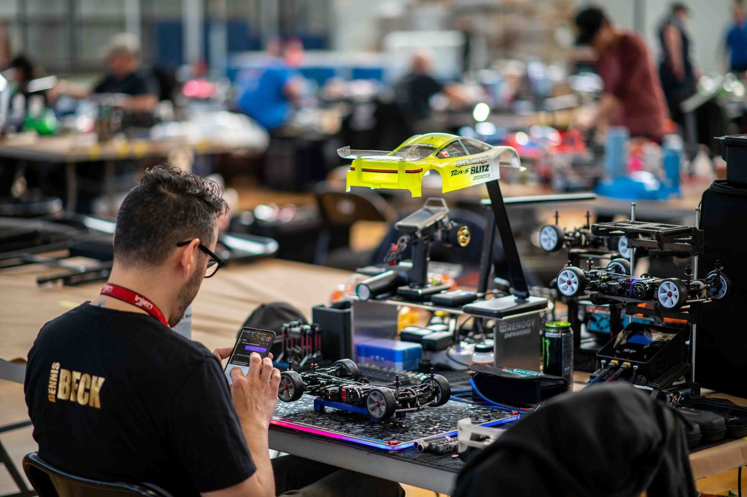 Man in black shirt assembling RC car at busy hobby racing competition.