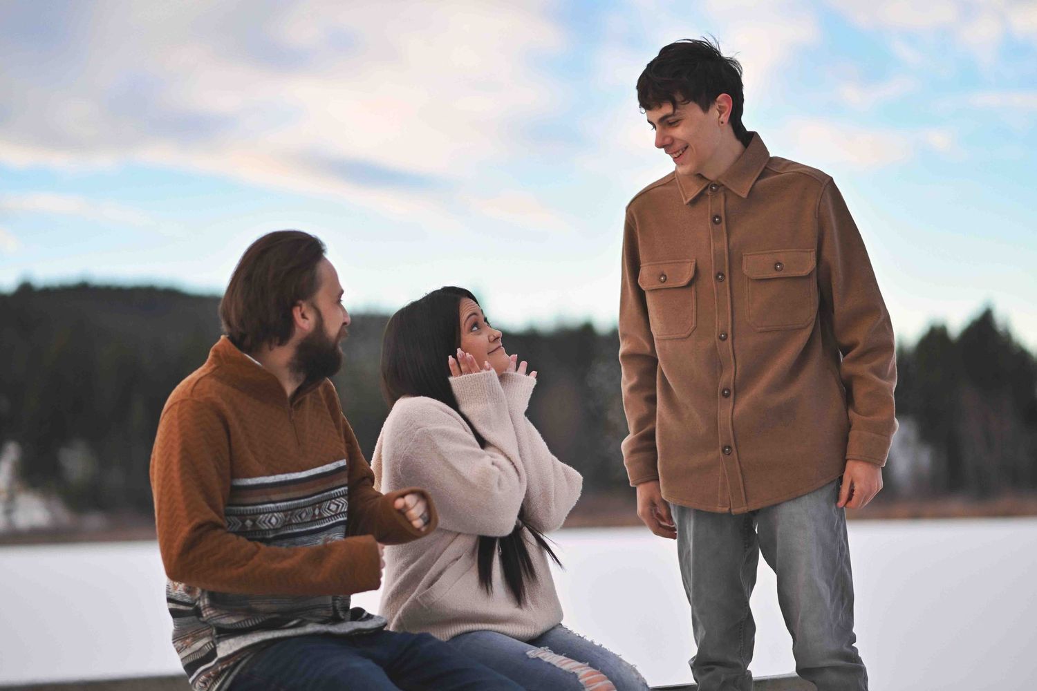 Three friends laugh together outdoors in winter near a frozen lake, wearing warm brown clothing.