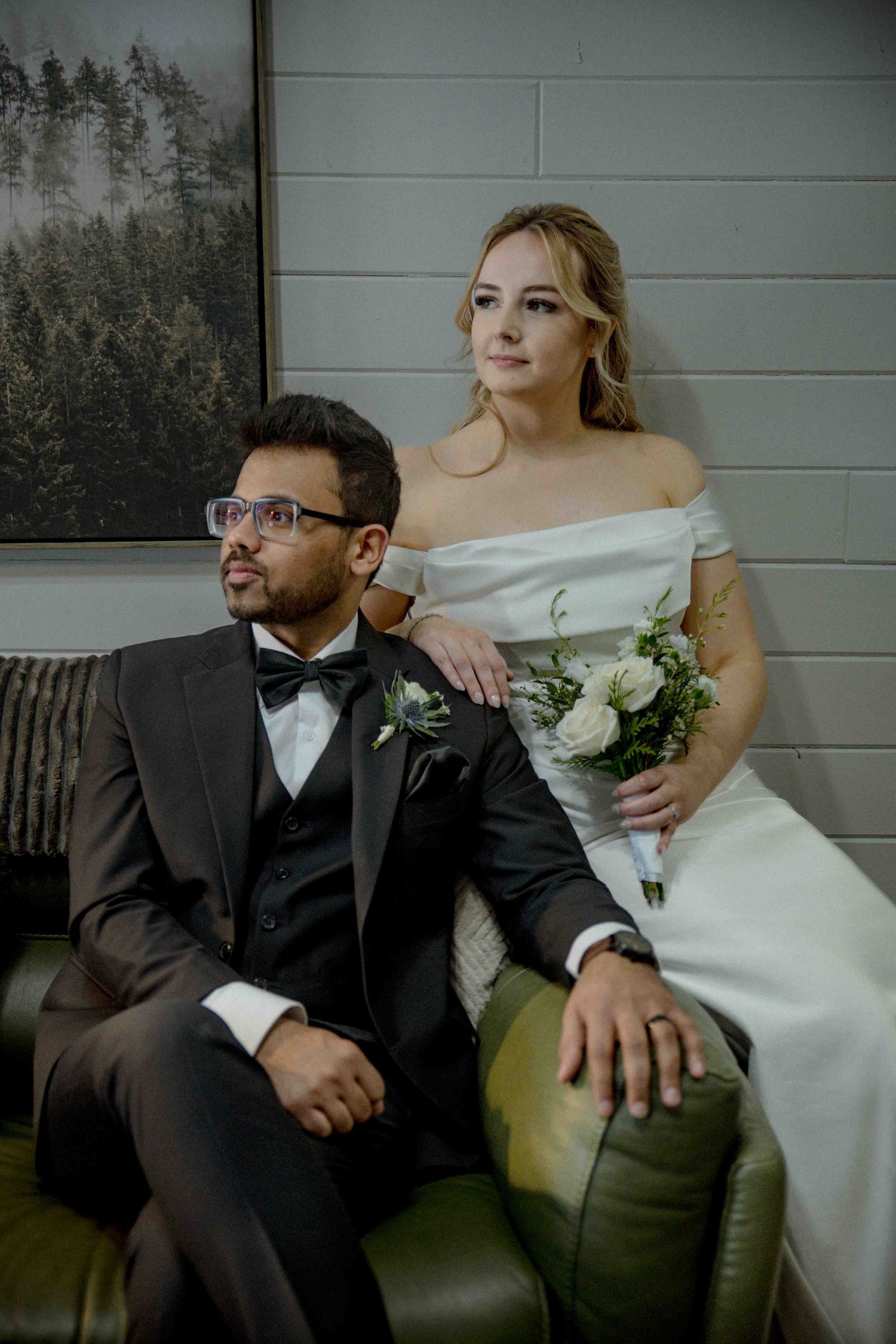 Bride and groom in formal wedding attire sitting on green leather sofa with white rose bouquet.